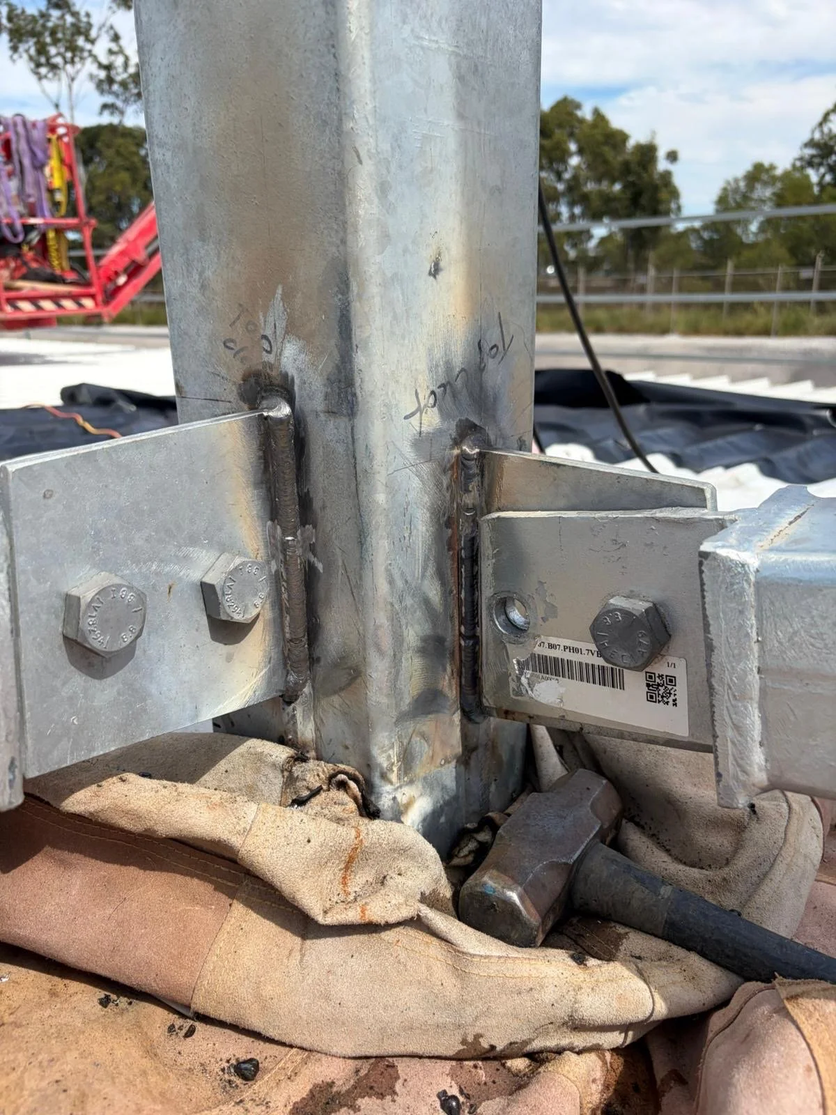 Close-up of a metal structure with bolts and welds, with handwritten notes and a barcode label, on a construction site or industrial setting.
