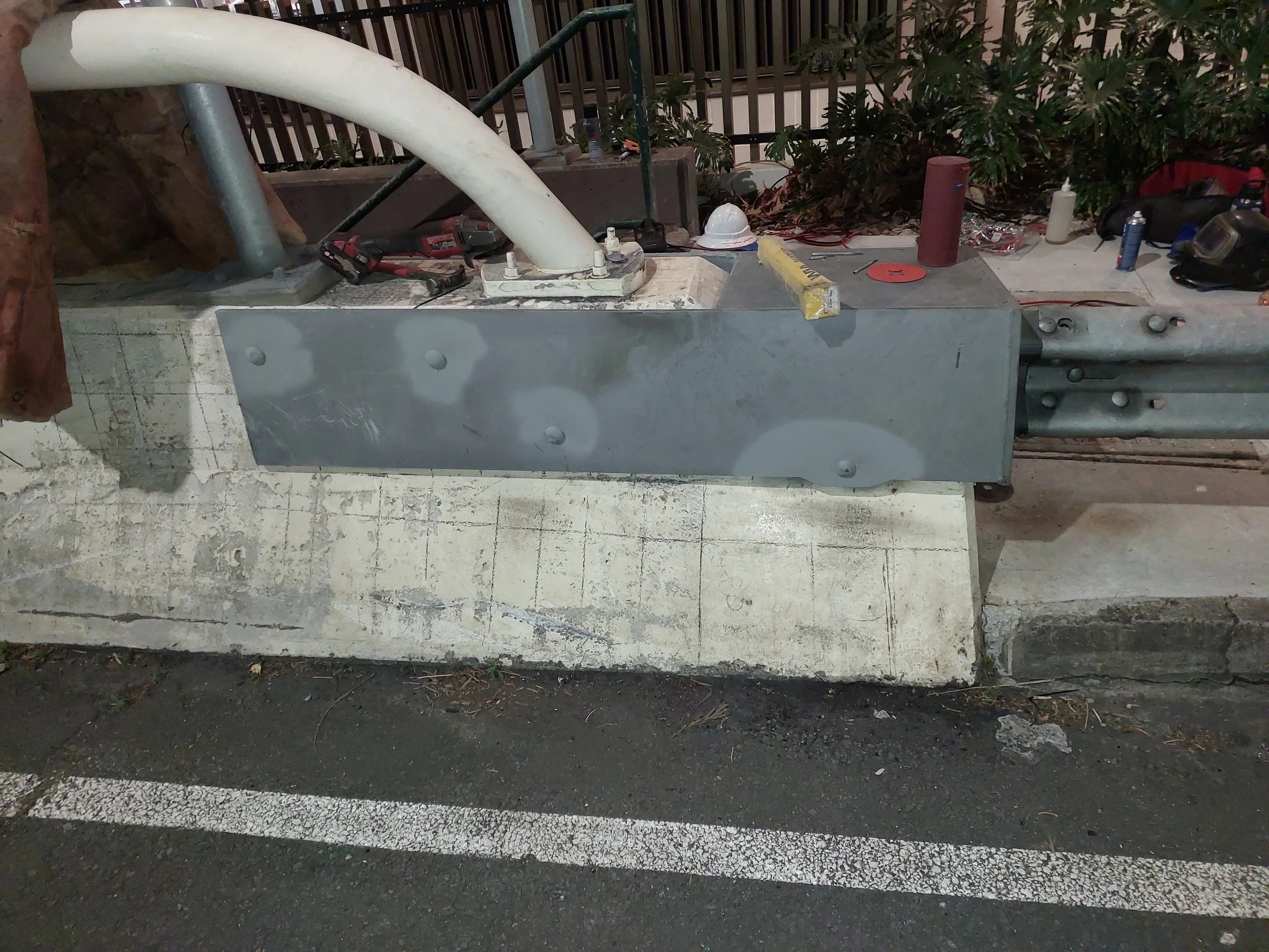 A construction area at night showing a metal guardrail, tools, construction materials, a hard hat, and surrounding foliage.