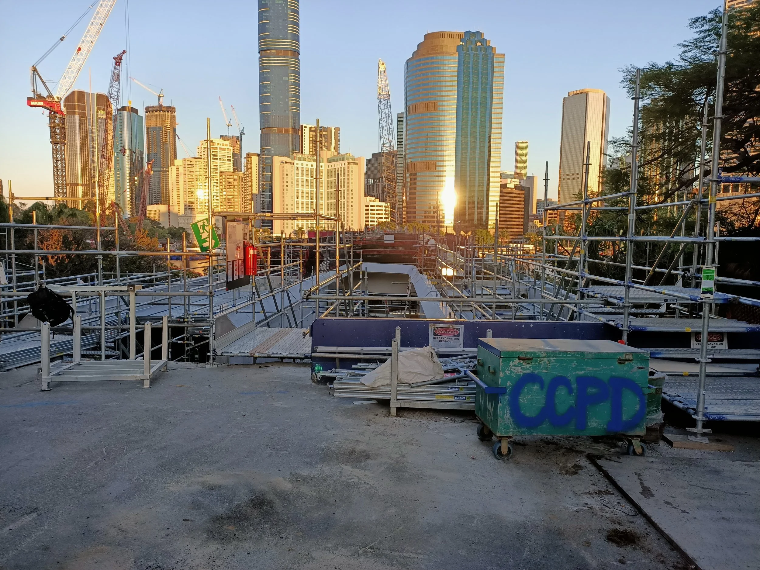 Construction site with metal scaffolding and equipment, and a city skyline with tall buildings in the background during sunset.