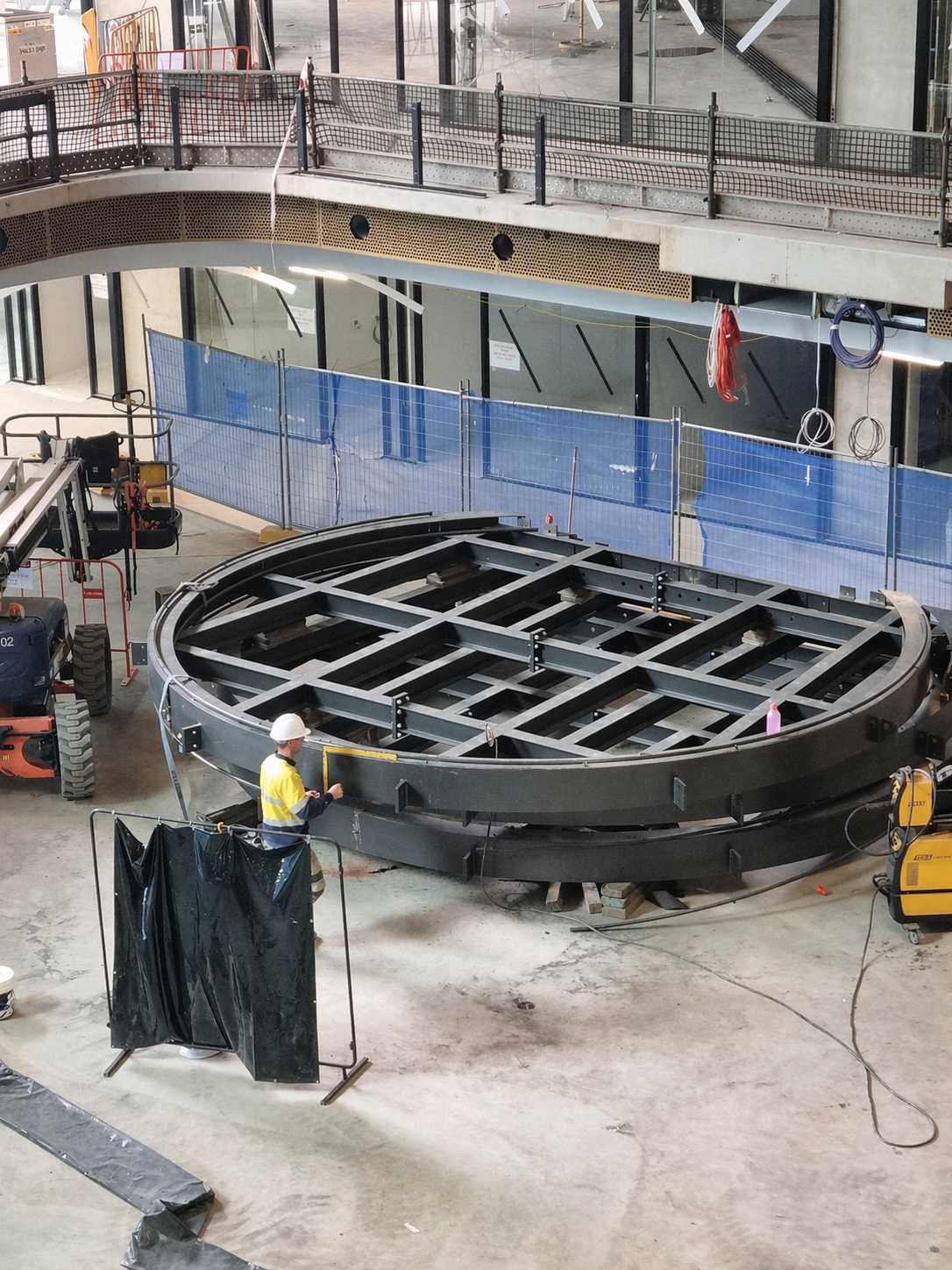 Construction site interior with a large circular metal structure being assembled, a worker in a yellow vest and white helmet working near the structure, scaffolding, tools, and construction barriers around the area.