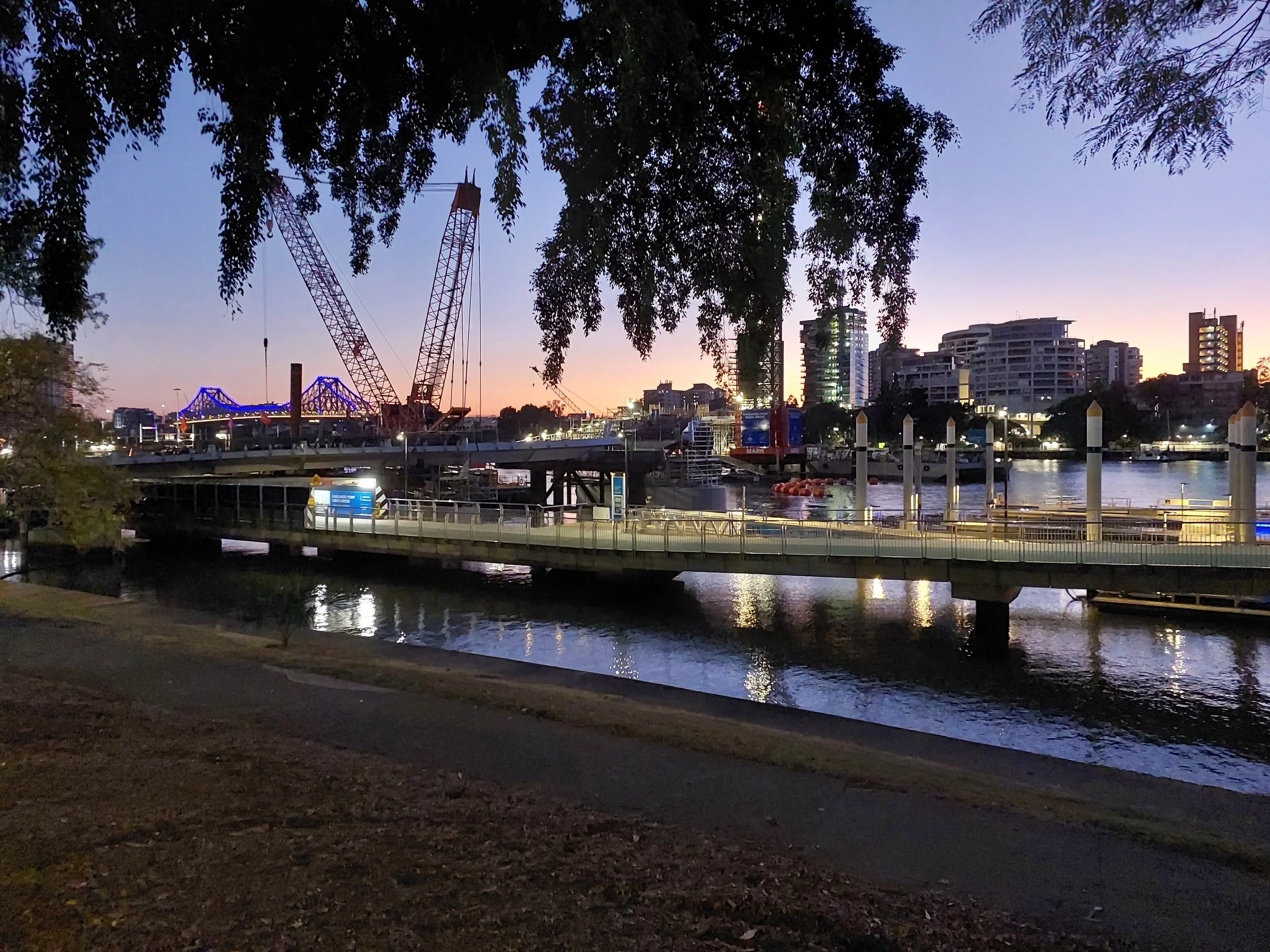A city waterfront at dusk with a bridge illuminated in blue, construction cranes, high-rise buildings, and a calm body of water reflecting the lights.