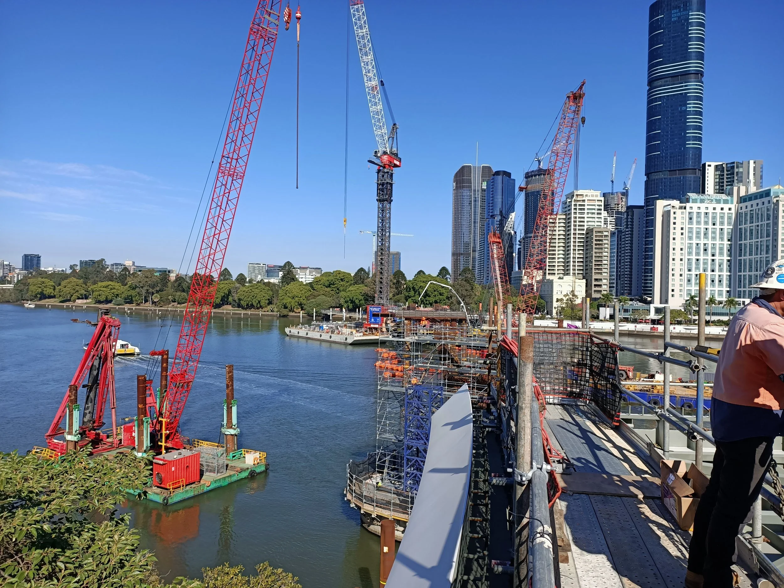 Construction site with cranes and scaffolding near a river, with tall buildings in the background.