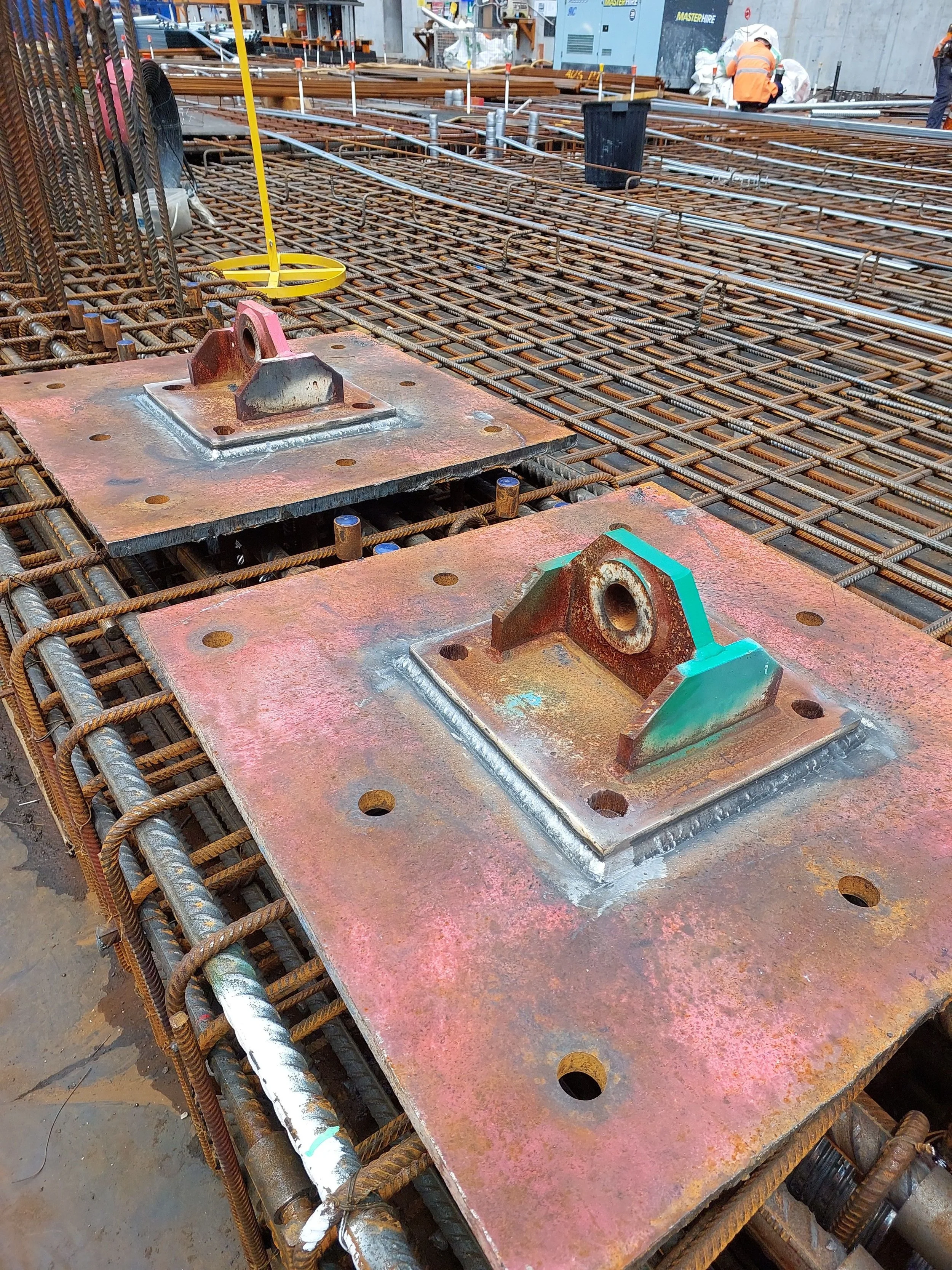 Construction site with reinforced steel bars forming a grid, steel plates with attached brackets, and workers in safety vests and hard hats working in the background.