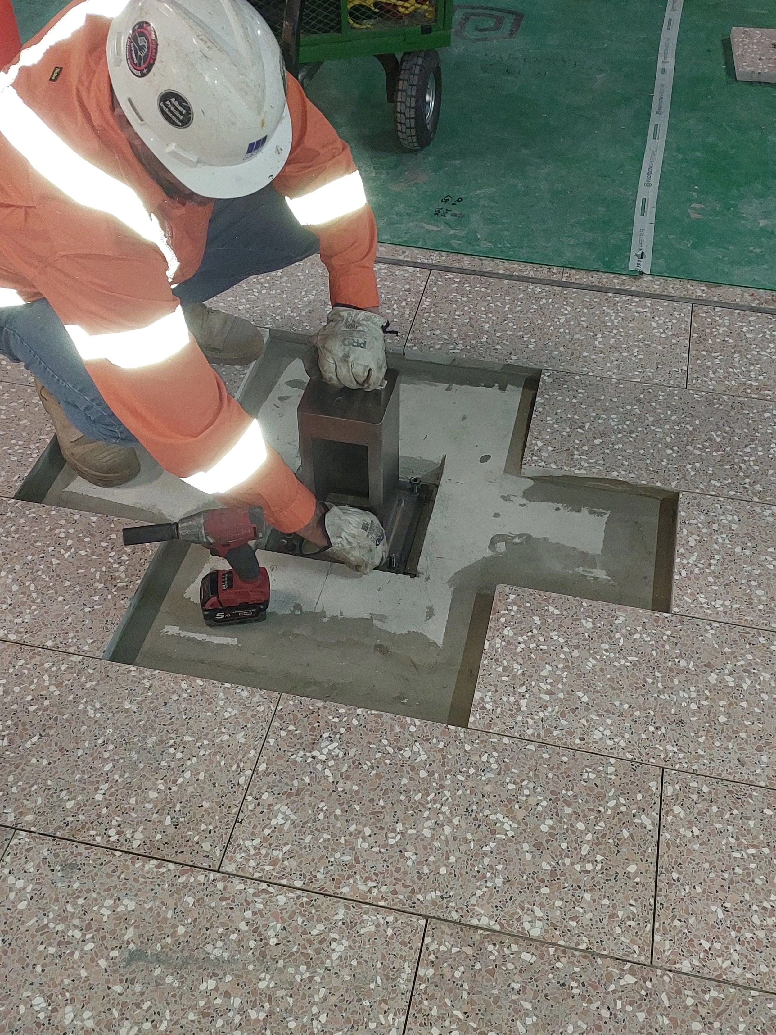 Construction worker installing tiles on the floor, wearing orange safety gear and a white helmet.
