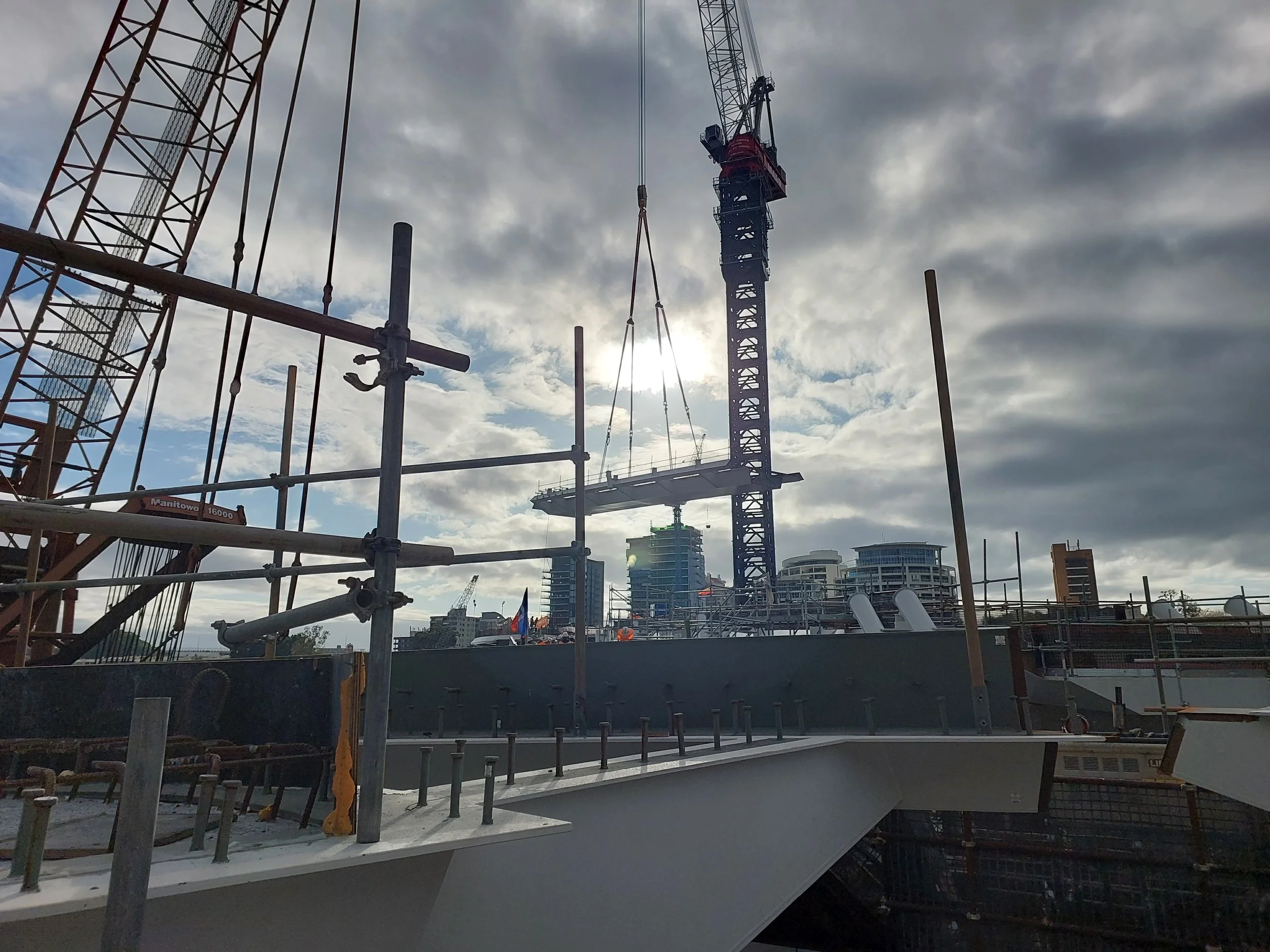 Construction site with scaffolding, cranes, and partial view of buildings in the background under cloudy sky.