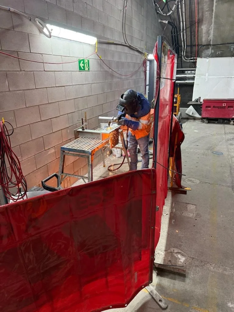 A construction worker wearing a helmet and safety gear welding metal on a worktable inside an industrial space with brick walls and fluorescent lighting fixtures.