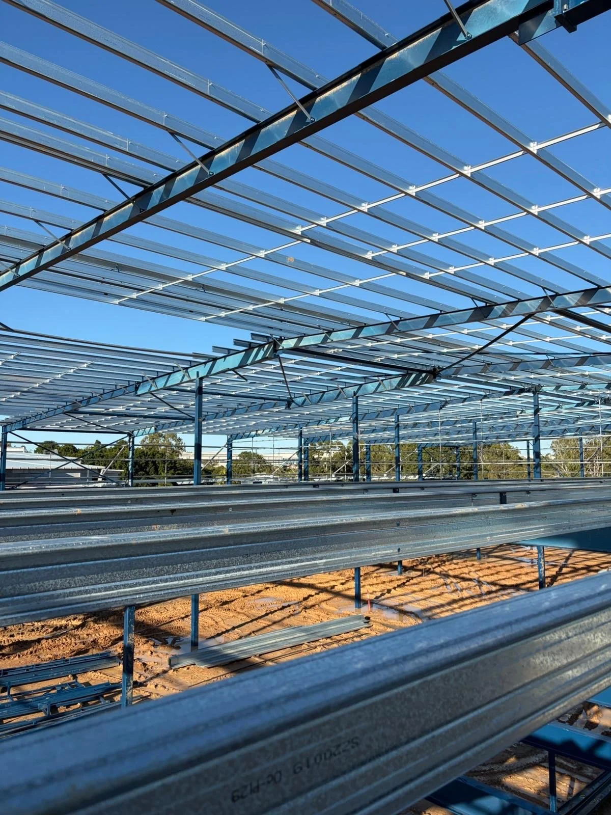 Steel structure of a solar panel installation on a construction site, with the blue sky in the background and trees visible beyond.