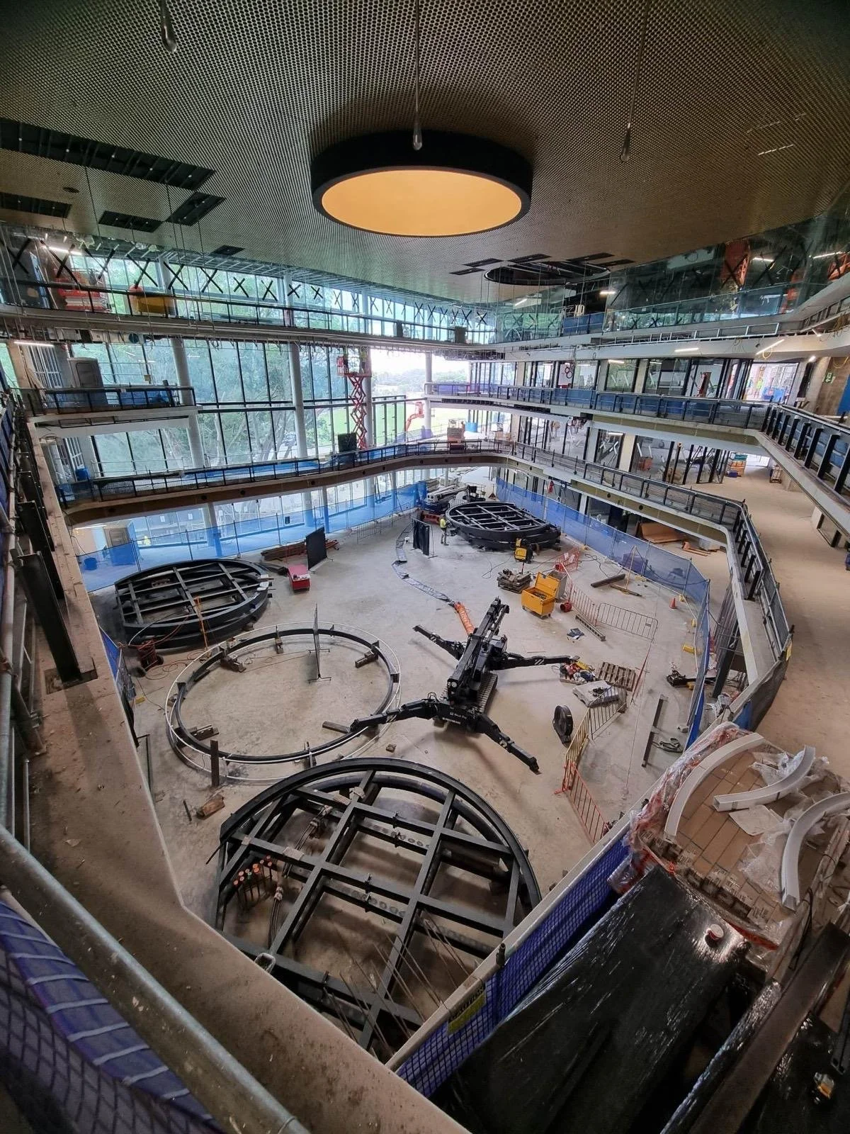 Interior under construction of a multi-level building with large glass windows, circular structures, construction equipment, and safety barriers.