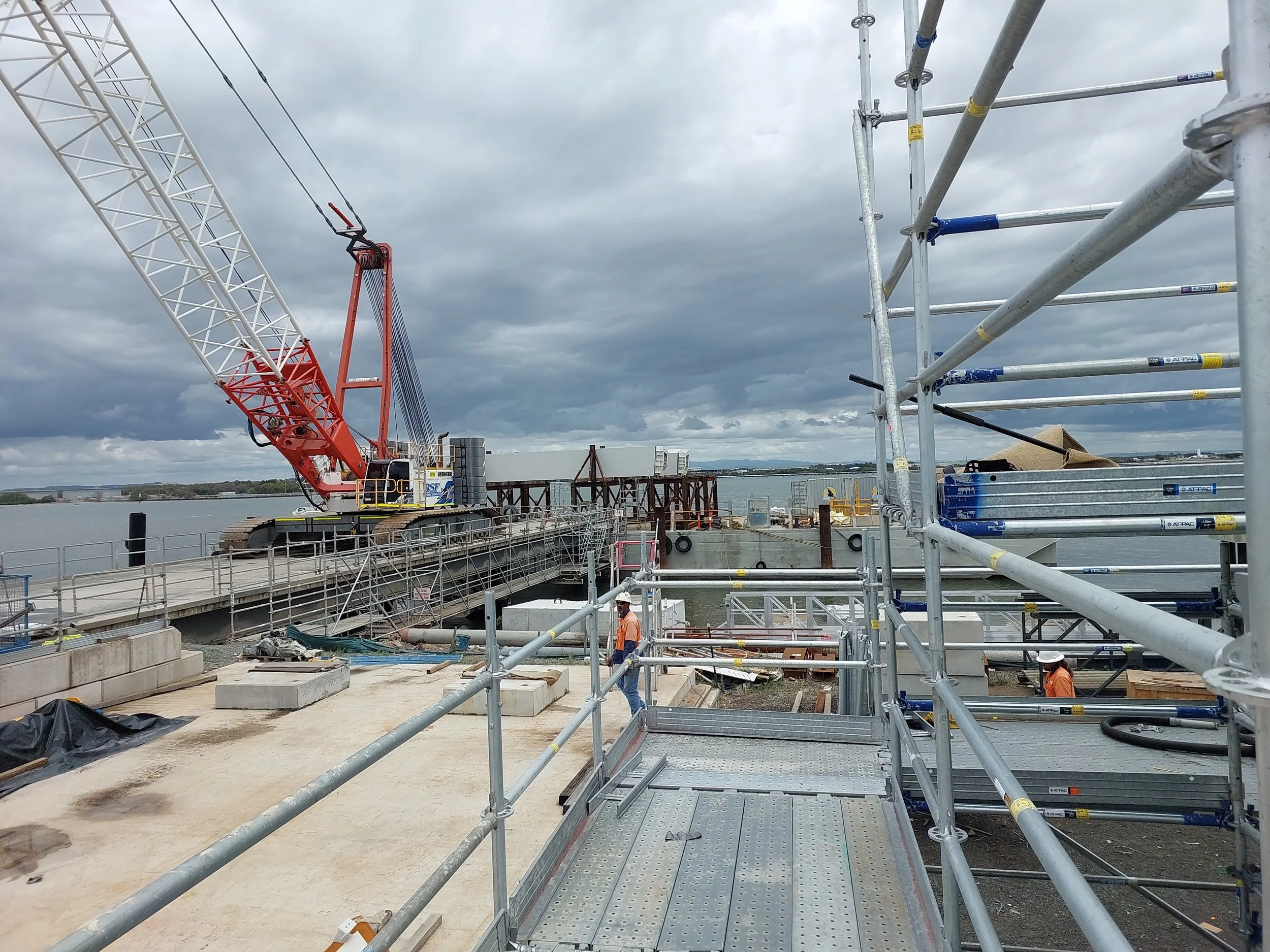 Construction site with scaffolding, a crane, and workers near a body of water under cloudy sky.