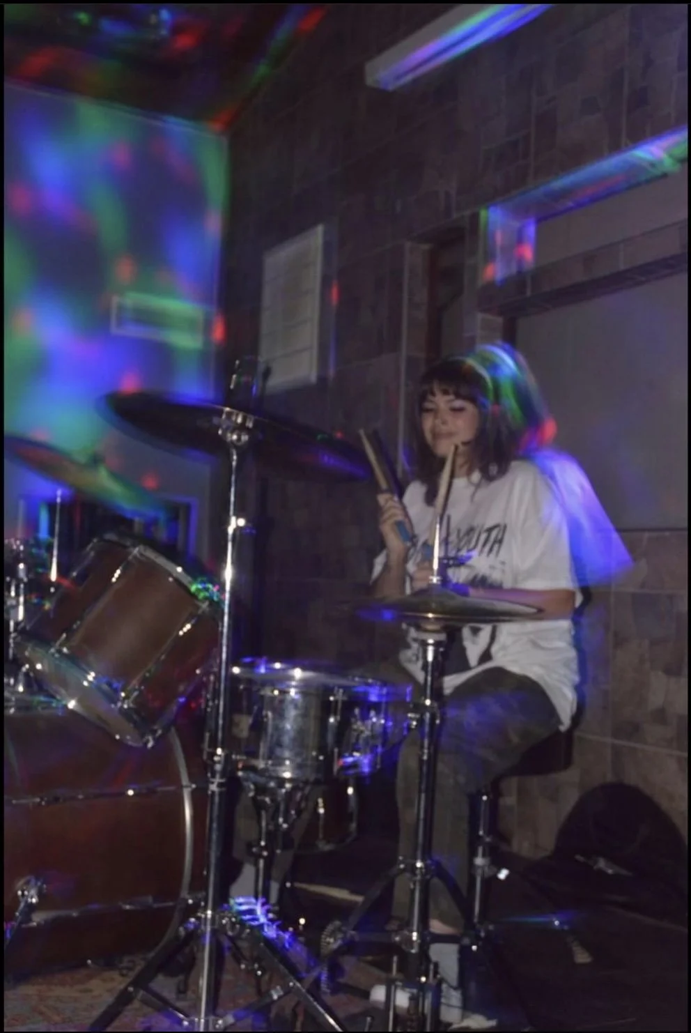 A young woman with headphones playing drums in a room decorated with colorful party lights.