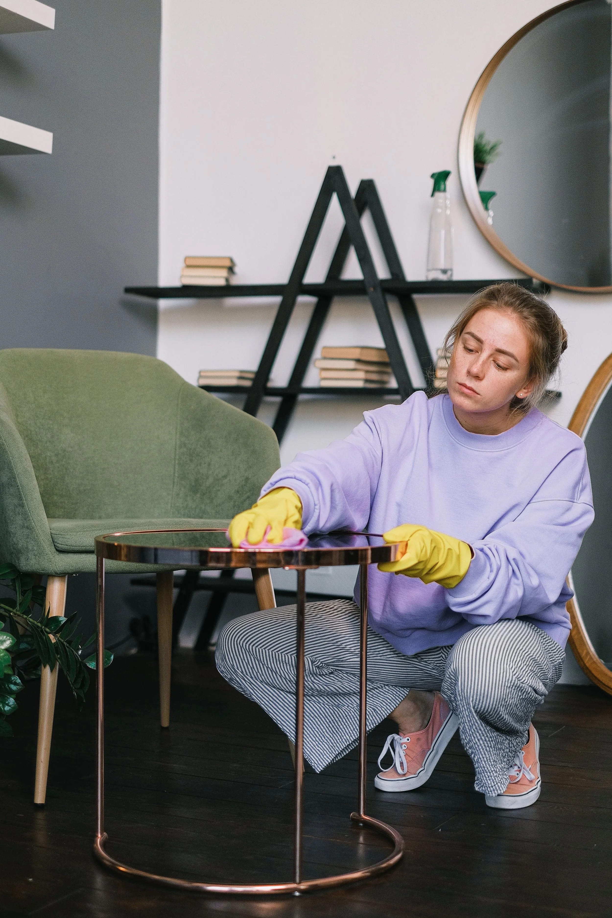 A young woman cleaning a copper-colored table with yellow gloves, wearing a lavender sweatshirt, striped pants, and pink sneakers, in a modern living room with a green armchair, black geometric bookshelf, and round mirror.