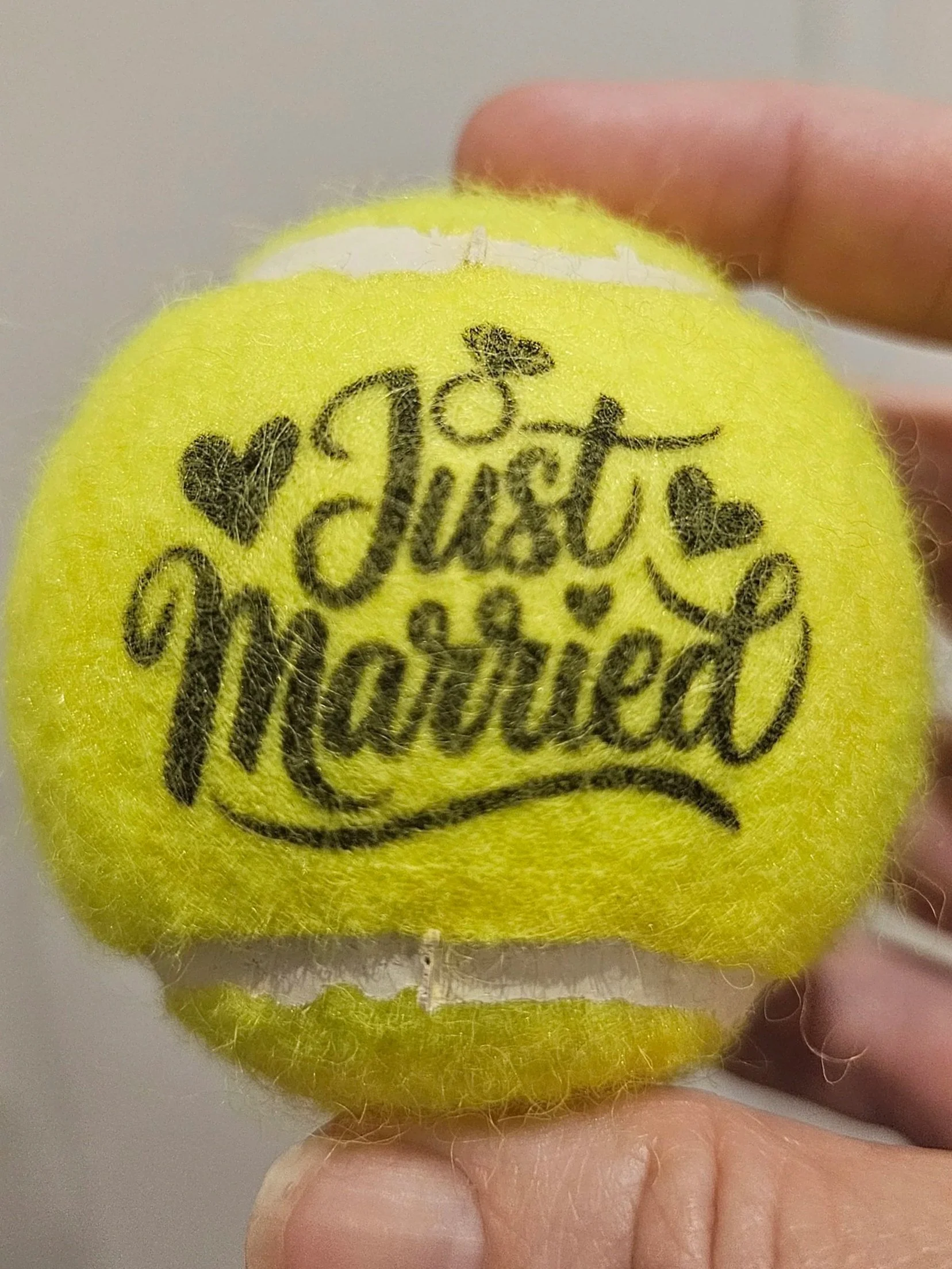 Close-up of a yellow tennis ball held between a person's thumb and index finger. The ball has black text and a small heart graphic on its surface. Wedding custom ball