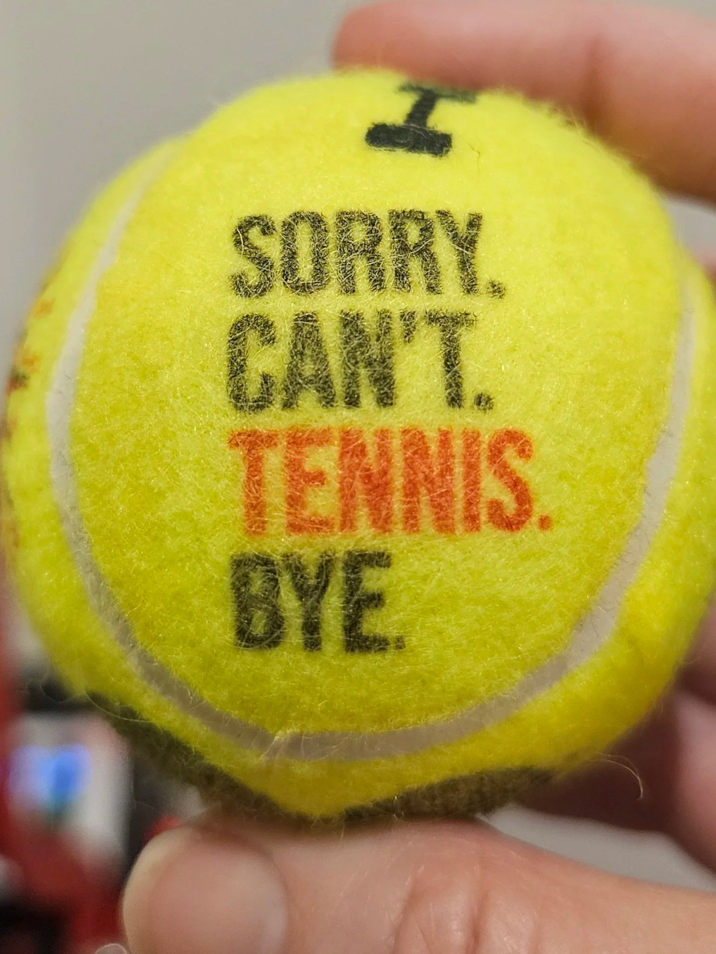 Close-up of a yellow tennis ball with black and red text. The ball is being held between fingers.