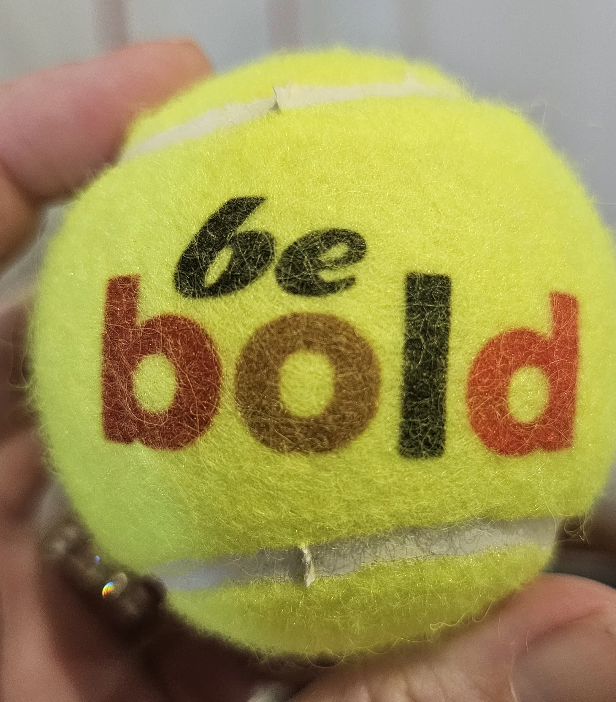 Close-up of a yellow tennis ball with black and red printed letters, held between person's fingers.