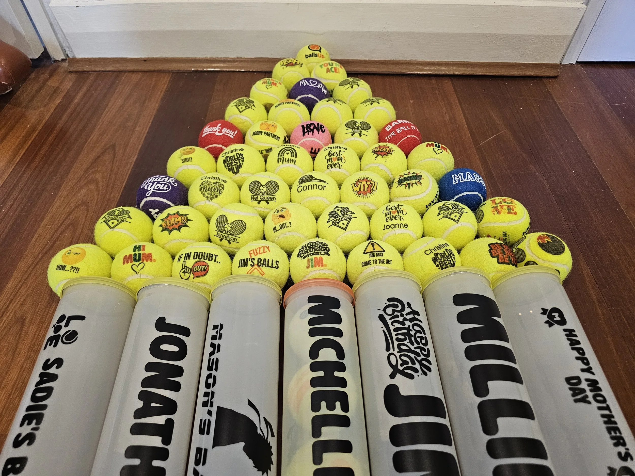 A pyramid of tennis balls decorated with various custom messages and graphics, sitting on a wooden floor against a wall.