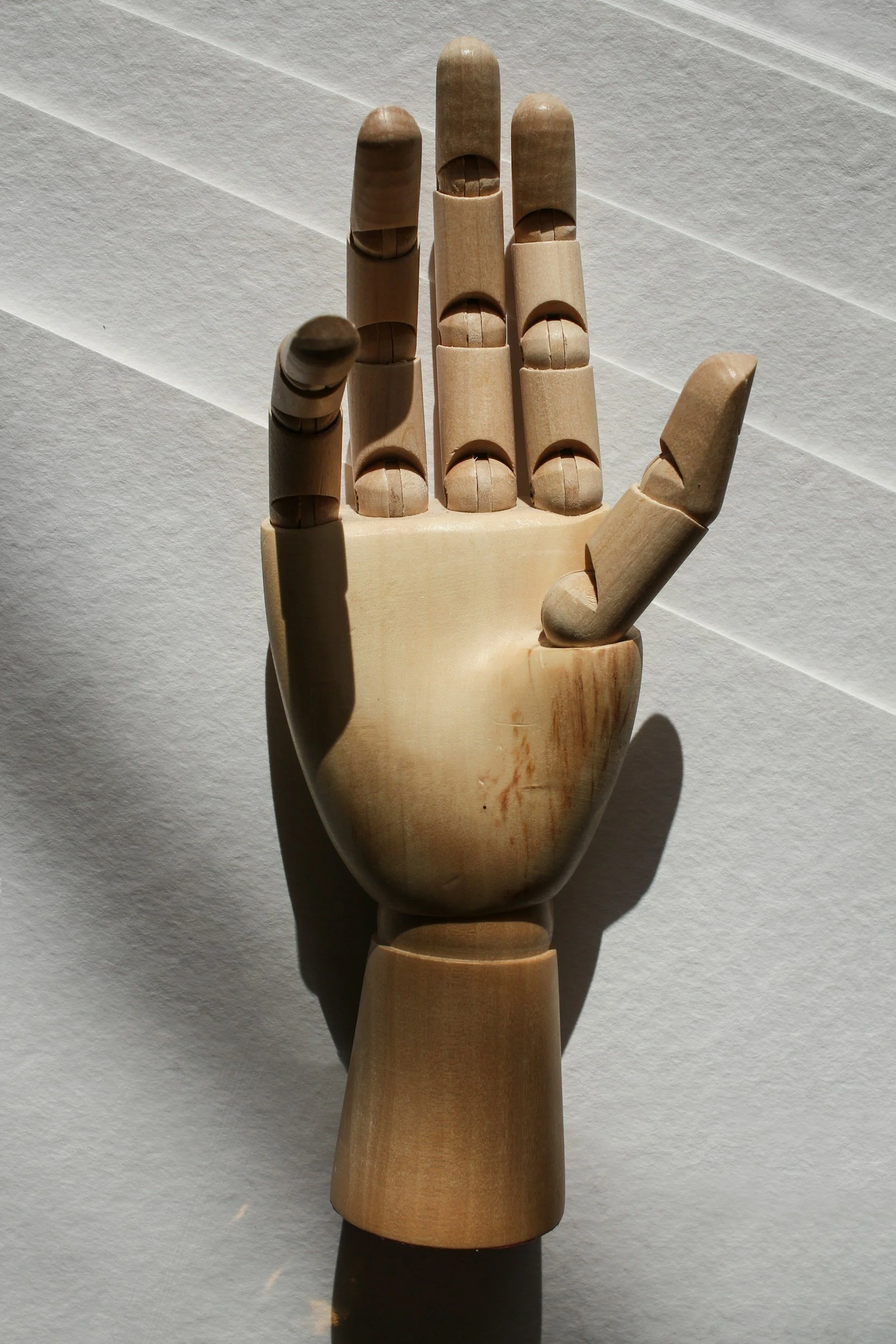 A wooden artist's mannequin hand in a palm-up position, with the fingers slightly spread apart, casting shadows on a white textured surface.