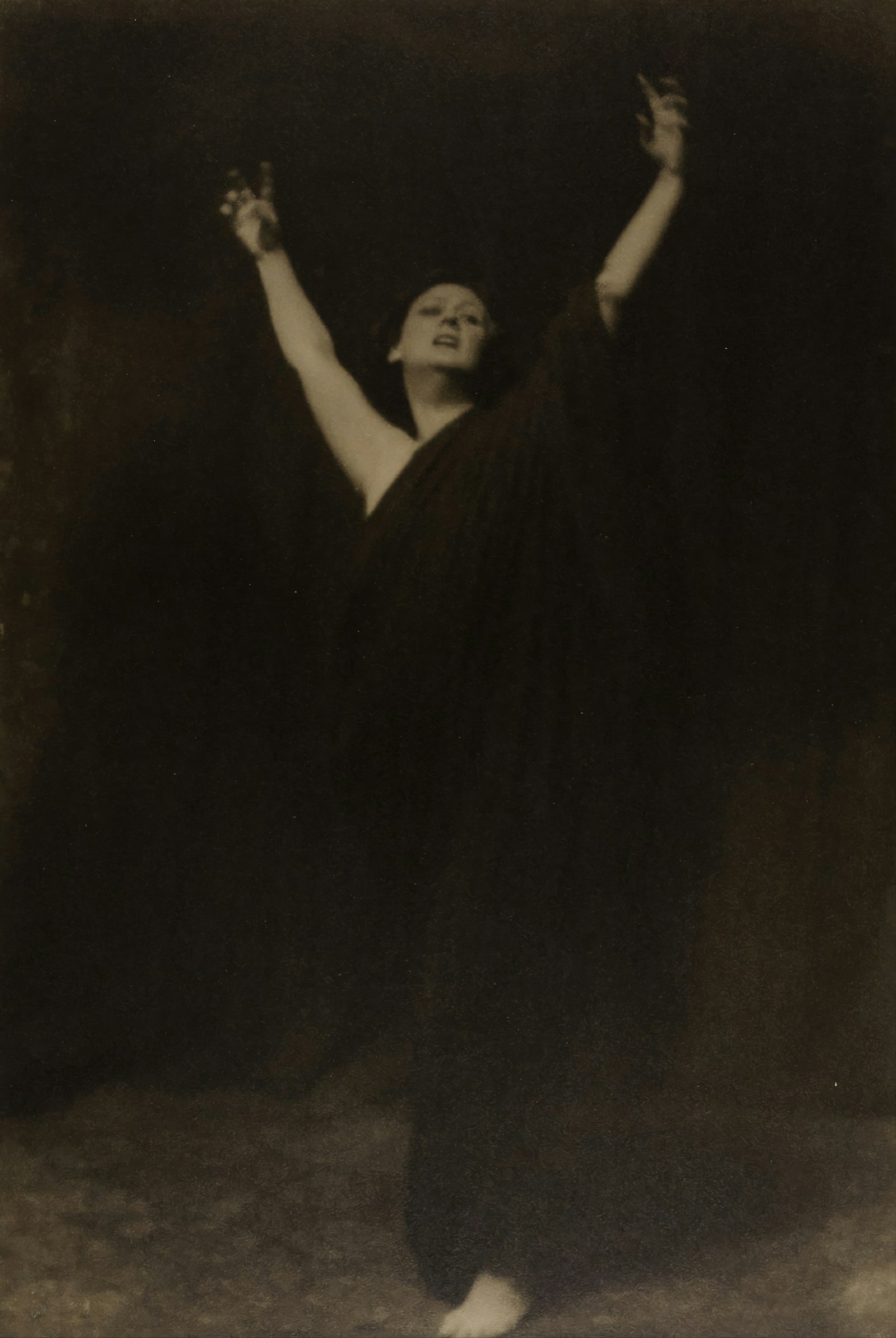 Black and white photo of a woman with arms raised, wearing a dark dress, in a dark setting.