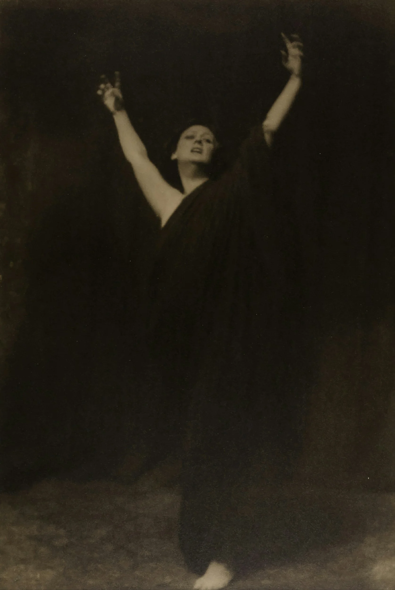 Black and white photo of a woman with arms raised, eyes closed, and head tilted back, wearing a dark dress.