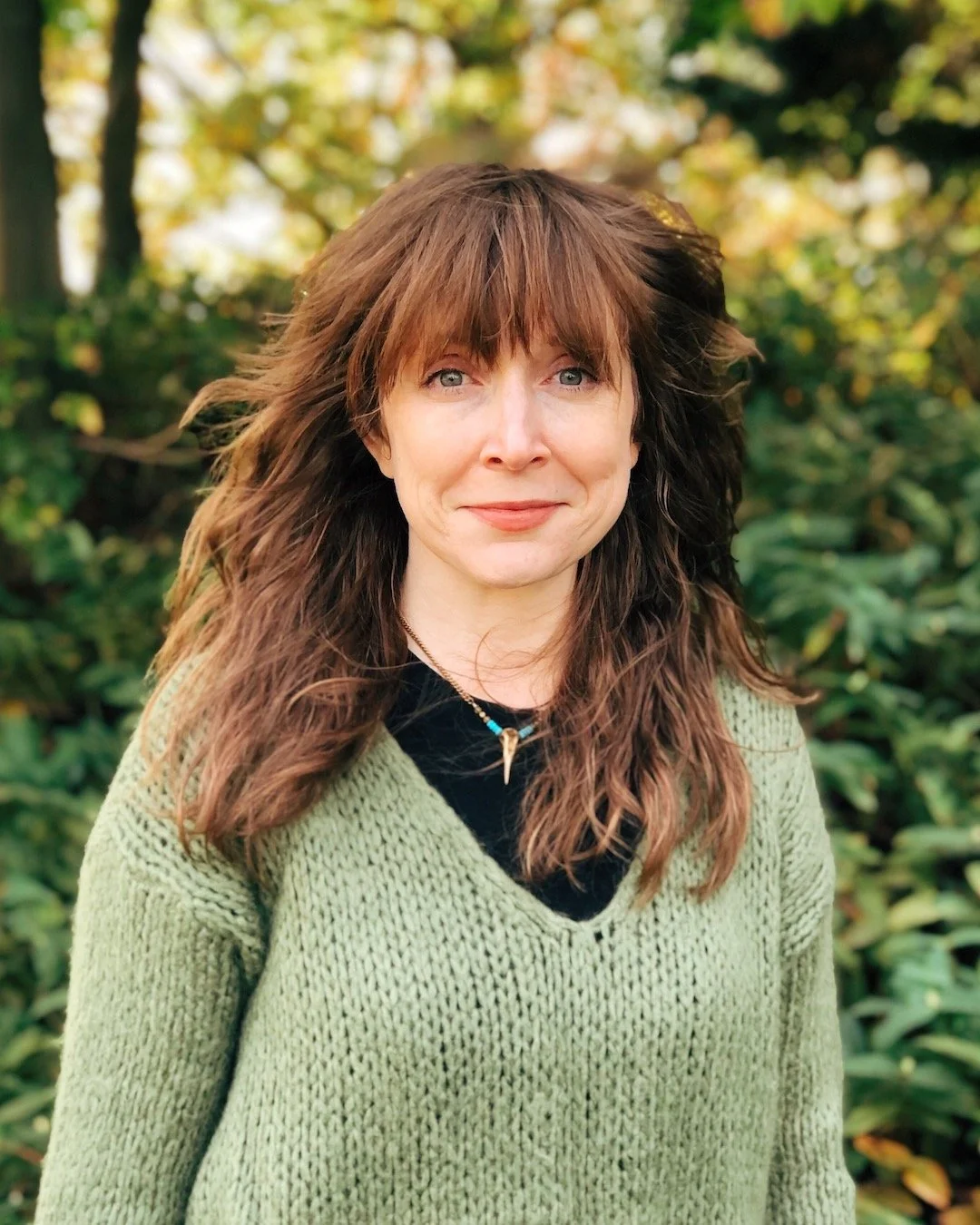 A woman with brown wavy hair and blue eyes outdoors with green foliage and trees in the background