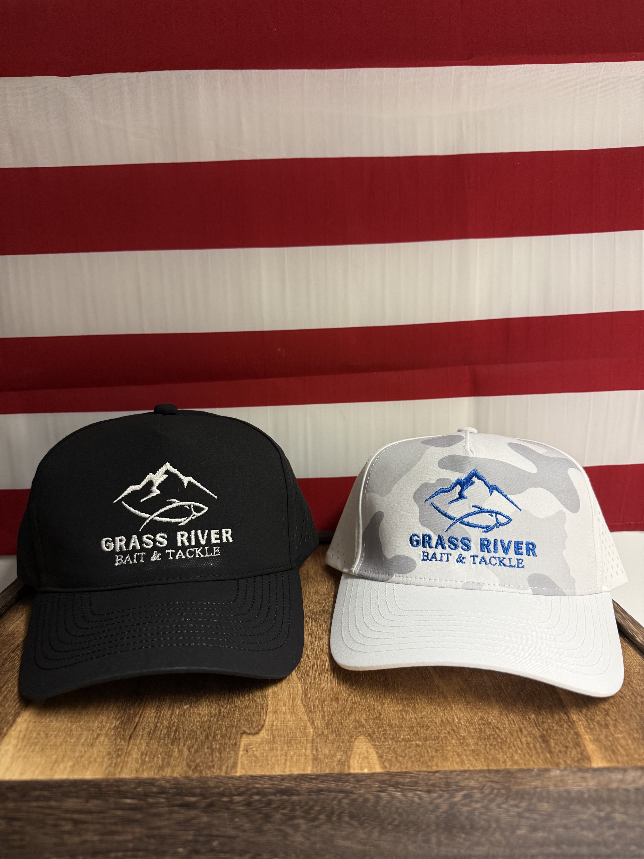 Two baseball caps, one black and one white, both embroidered with a mountain and fish logo and the words "Grass River Bait & Tackle", placed on a wooden table with an American flag hanging in the background.