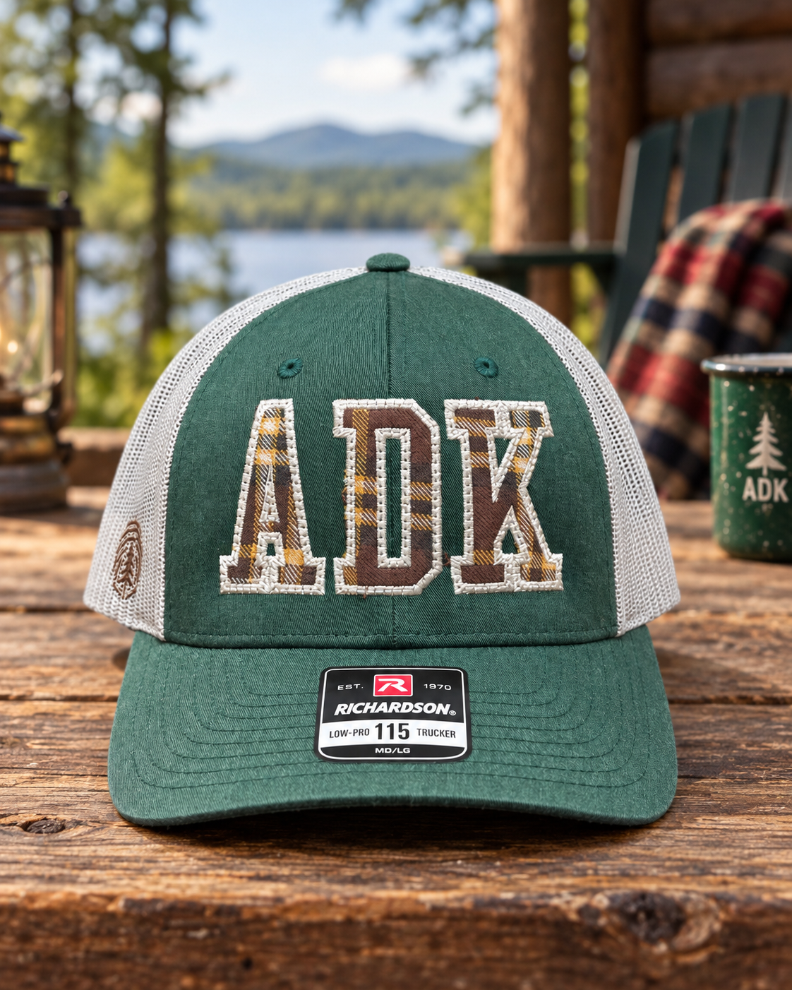 Green trucker hat with ADK in plaid pattern embroidered on the front, placed on a wooden table outdoors with a lake and trees in the background.
