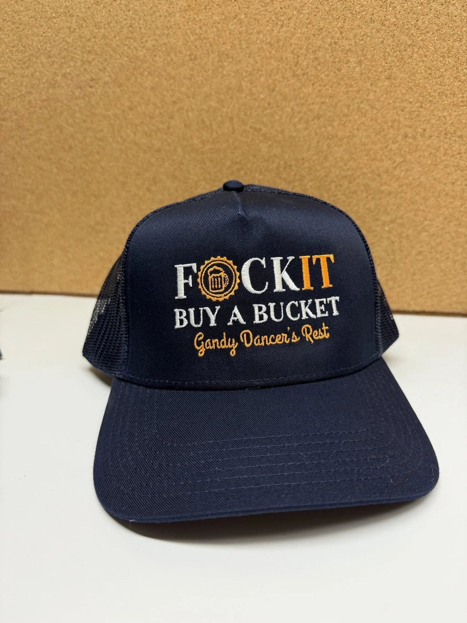 A navy blue baseball cap with embroidered text that reads 'F**K IT BUY A BUCKET Gandy Dancer's Rest' in white and orange letters, placed on a white surface against a brown background.