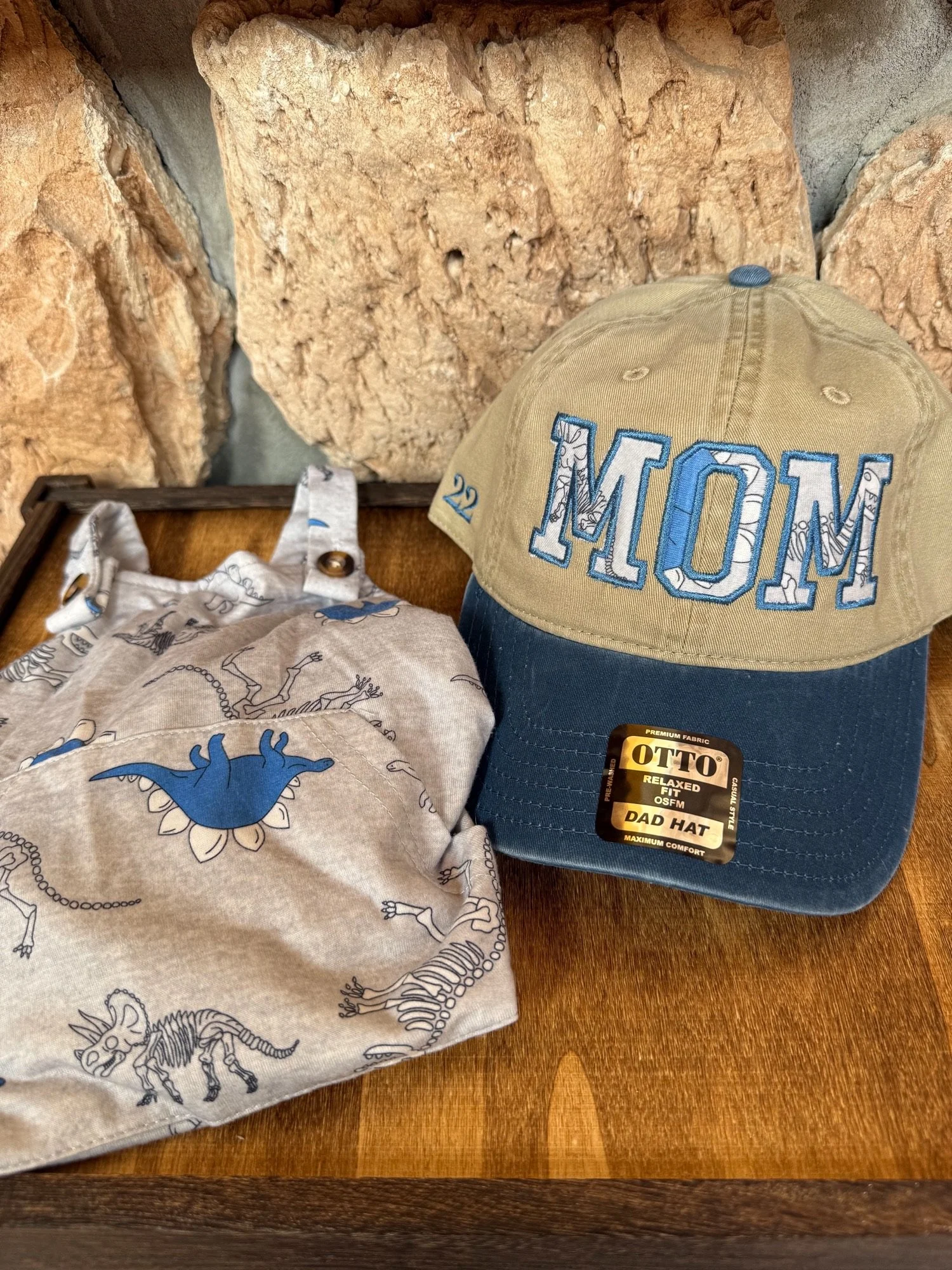 A beige and navy blue dad hat with 'MOM' embroidered in large blue and white letters, placed on a wooden surface. The hat has a tag that says 'Otto Relaxed Fit OSFM Dad Hat.' Next to the hat, there is a cloth with dinosaur and skeleton prints, and a fabric bag with a dinosaur design. The background shows a wall made of large, textured sand-colored rocks.