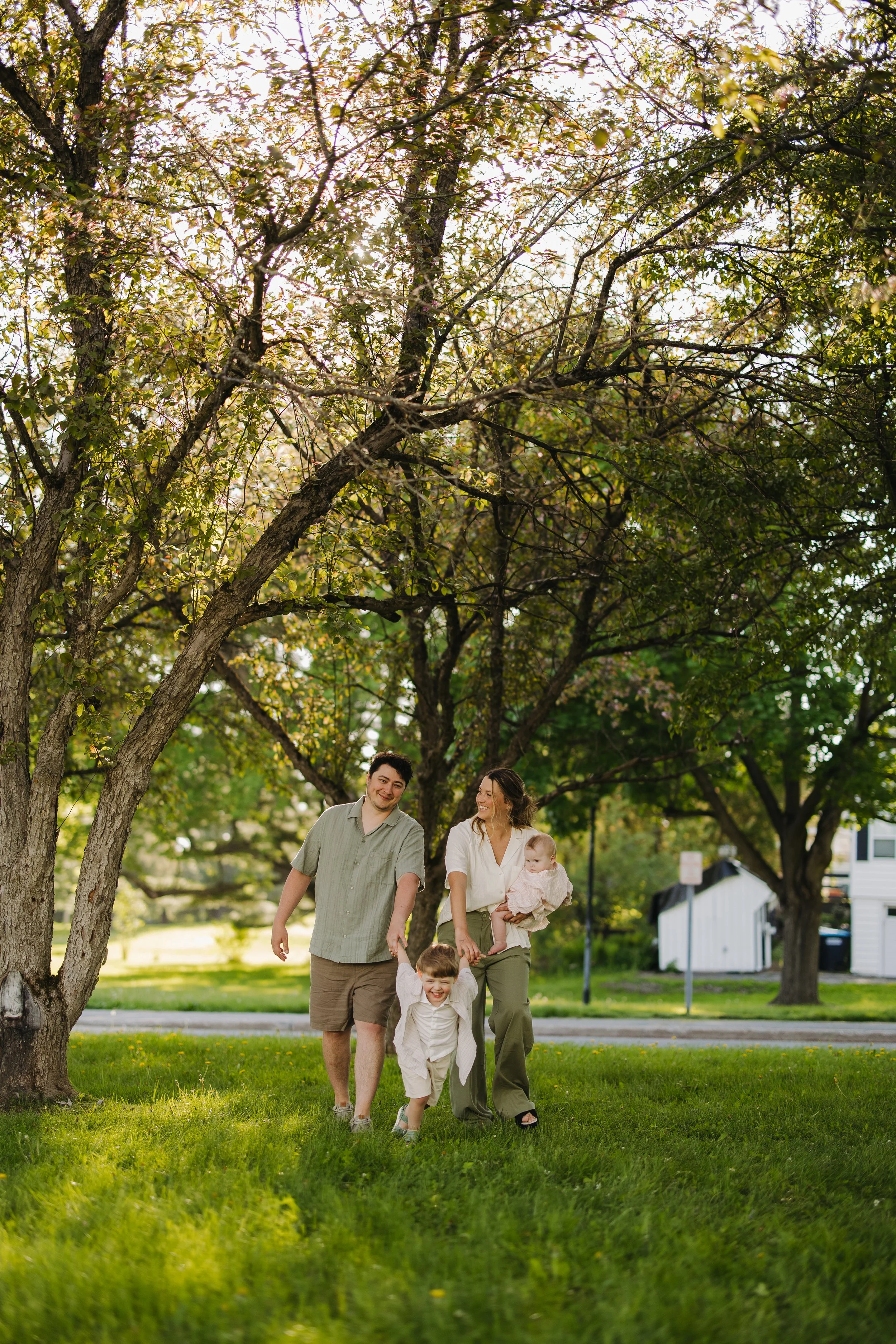 A family of four walking through a park on a sunny day. The trees have green leaves, and there is a house and street in the background.
