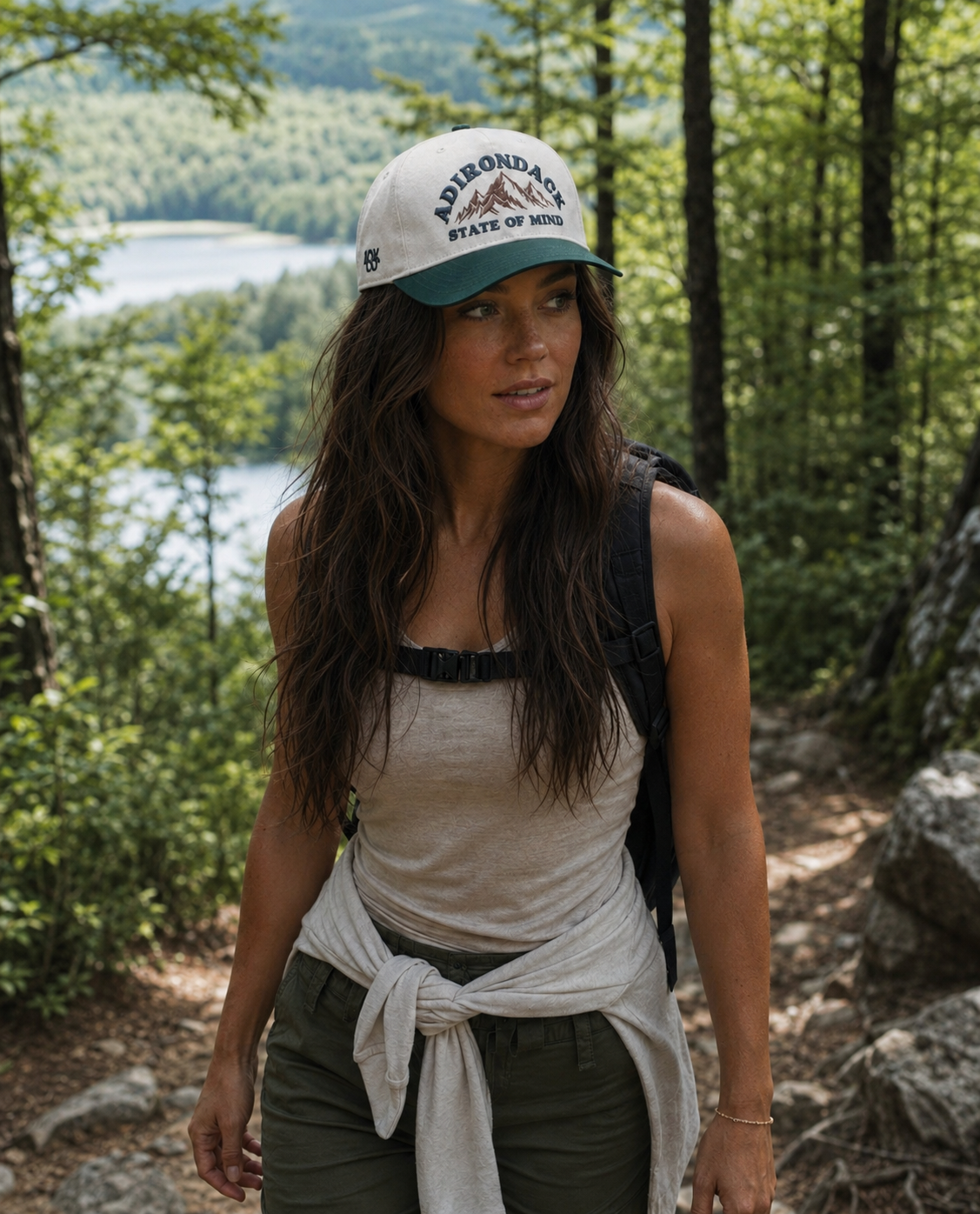 A woman with long brown hair, wearing a white and green cap, beige tank top, green pants, and a black backpack, hiking on a forest trail with trees and a lake in the background.