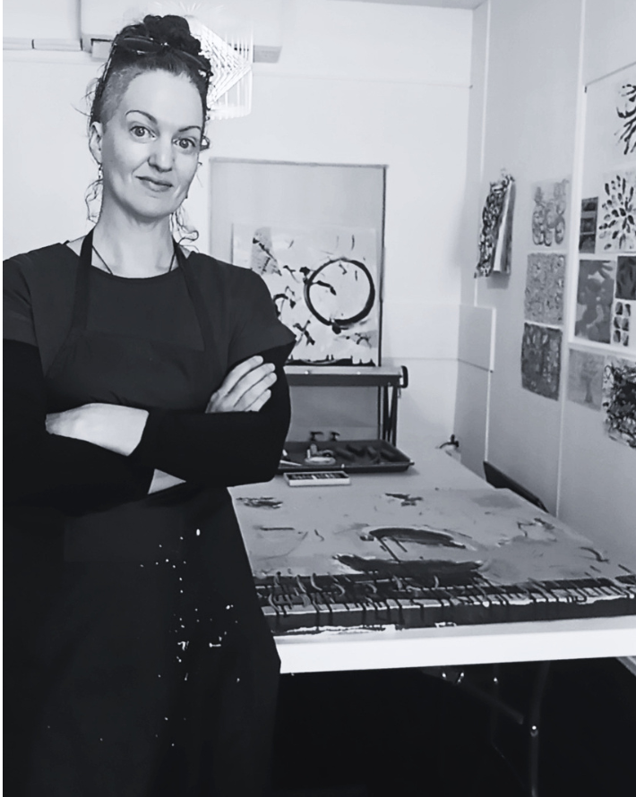 A woman with dark hair in a bun, dressed in black, standing with arms crossed in an art studio. In the background, there are abstract paintings, a table with art supplies, and artwork displayed on the walls.