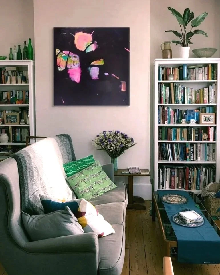 Living room with a beige sofa, colorful pillows, two white bookshelves filled with books, a wall-mounted abstract painting, a side table with a bouquet of purple flowers, a wooden coffee table with decorative plates, and a potted plant on top of the bookshelf.