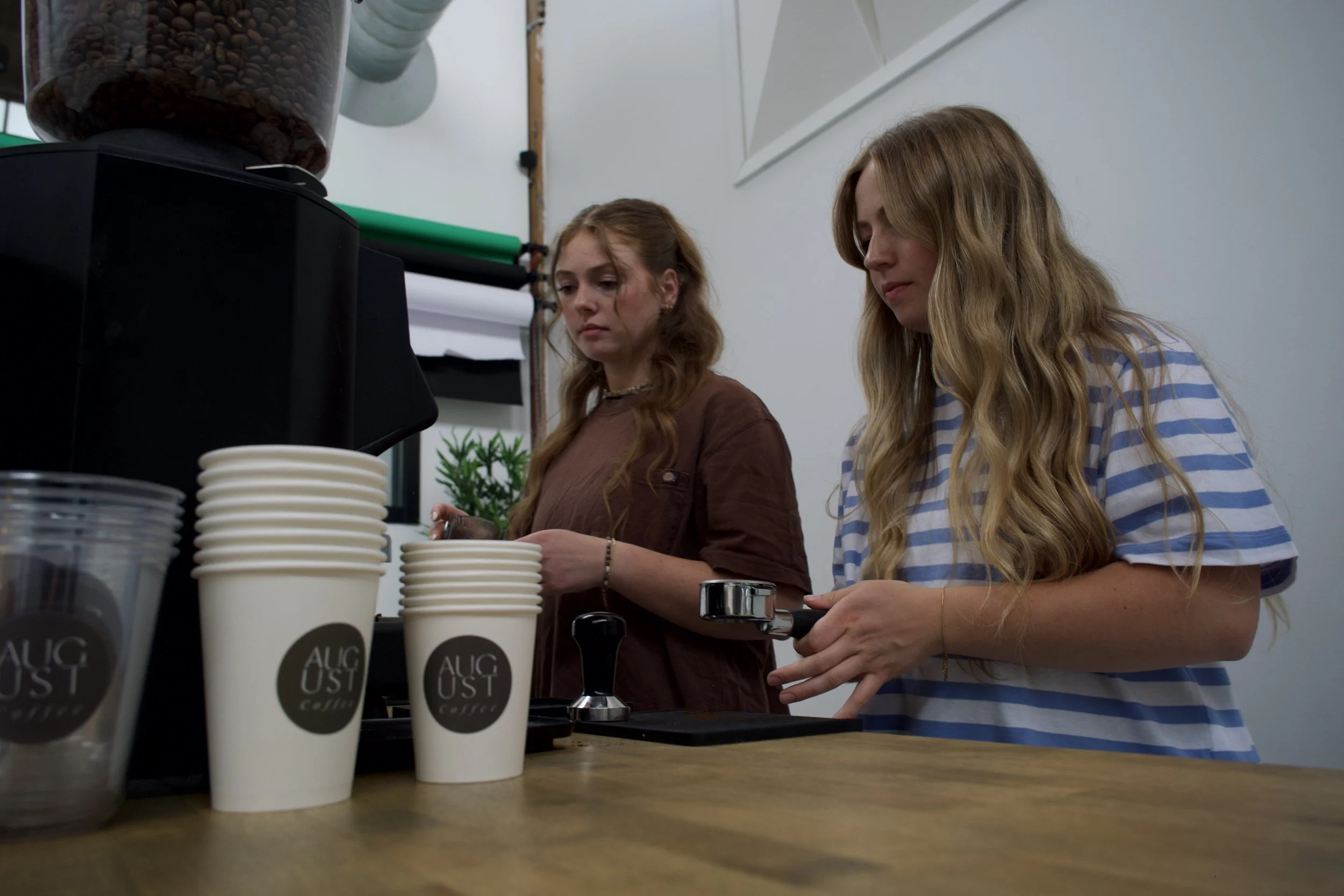 Two young women are standing at a coffee counter, preparing drinks. One has long blonde hair and is wearing a blue and white striped shirt, while the other has reddish-brown hair and is wearing a brown shirt. Coffee cups labeled 'AUGUST Coffee' are stacked on the counter.