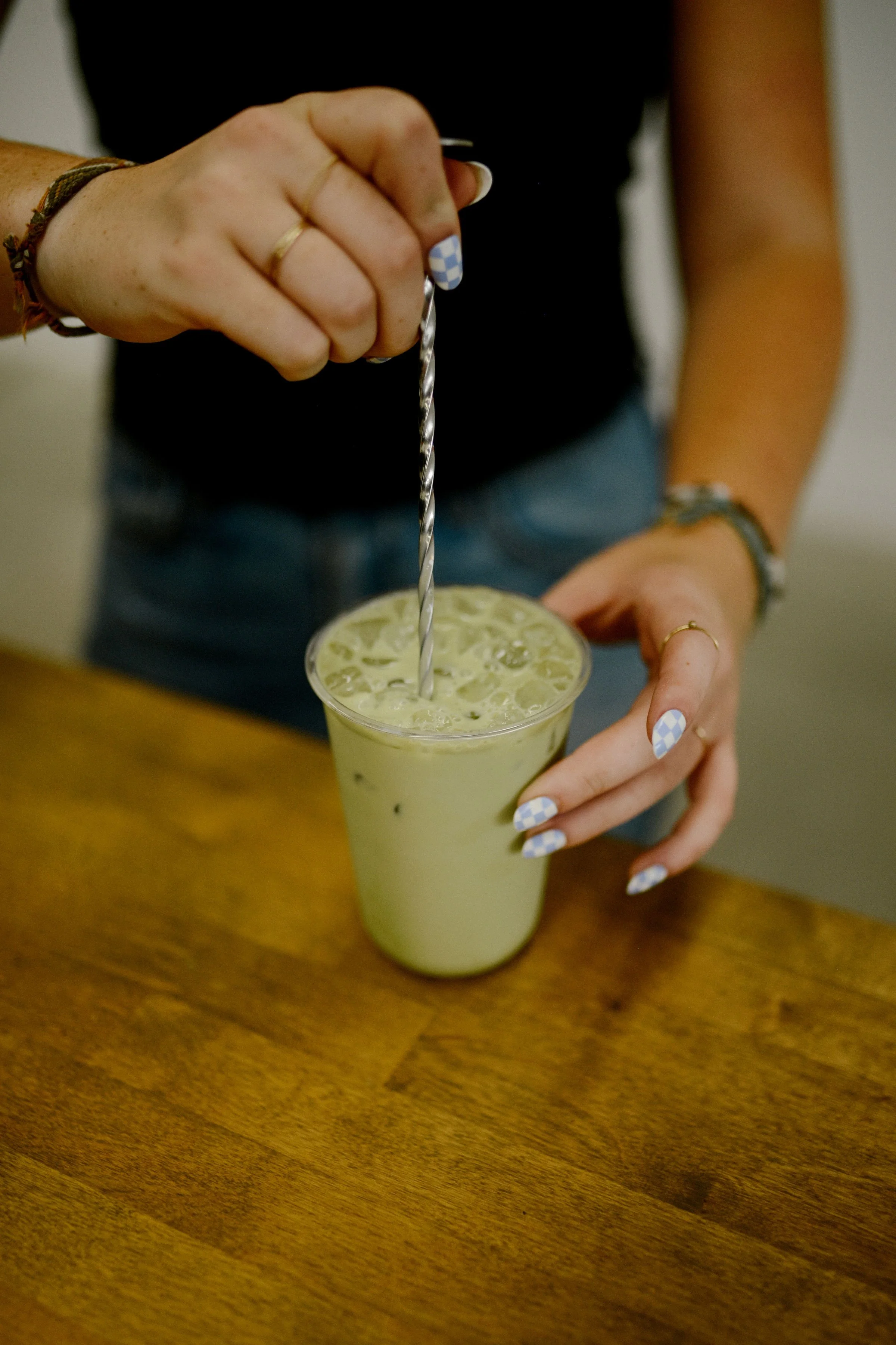 Person holding a glass with a light green beverage, ice, and a spiral straw, with painted nails and jewelry.