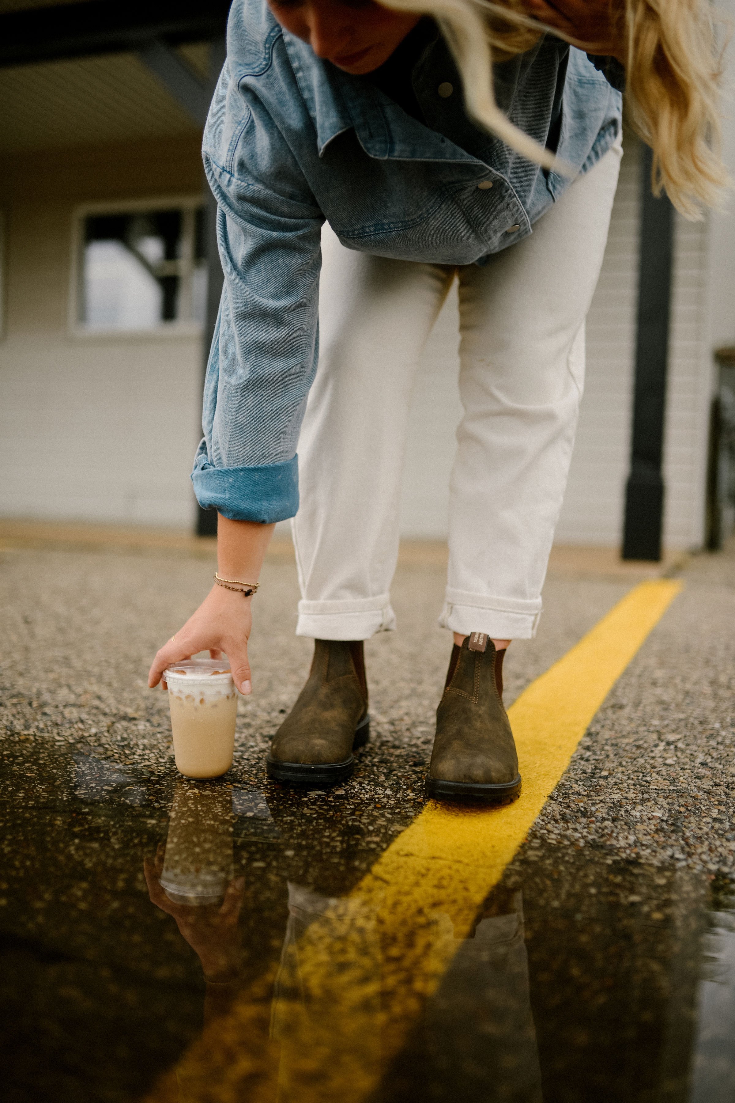 Woman in denim jacket and white jeans reaching for iced coffee on wet parking lot with yellow line.