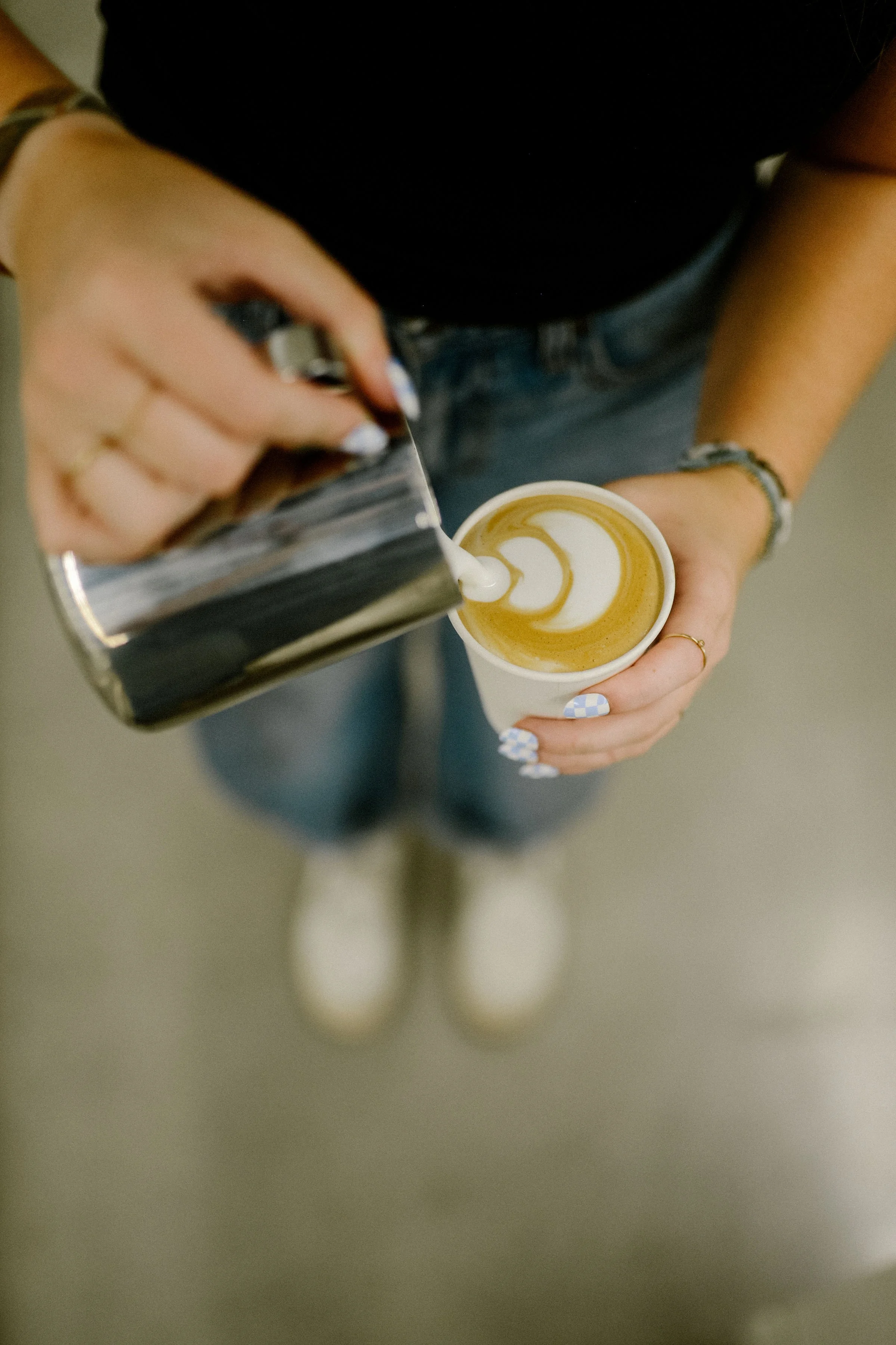 A person pouring milk into a cup of coffee with latte art on top, viewed from above. The person is wearing a black shirt, jeans, and has bracelets and rings on their hands.