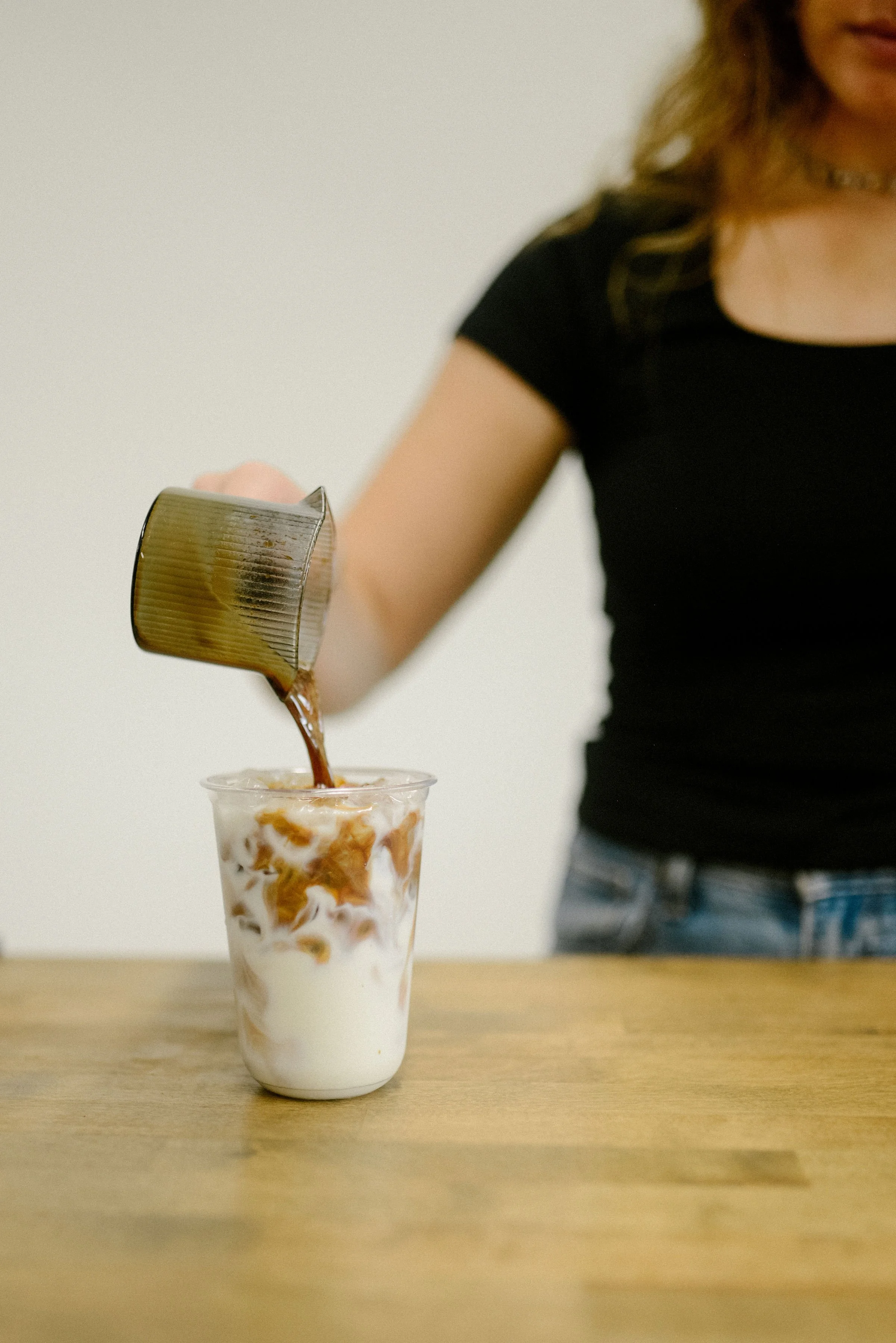 A woman in a black shirt and blue jeans pours a shot of organic espresso into a clear to go cup with milk and ice on a wood counter