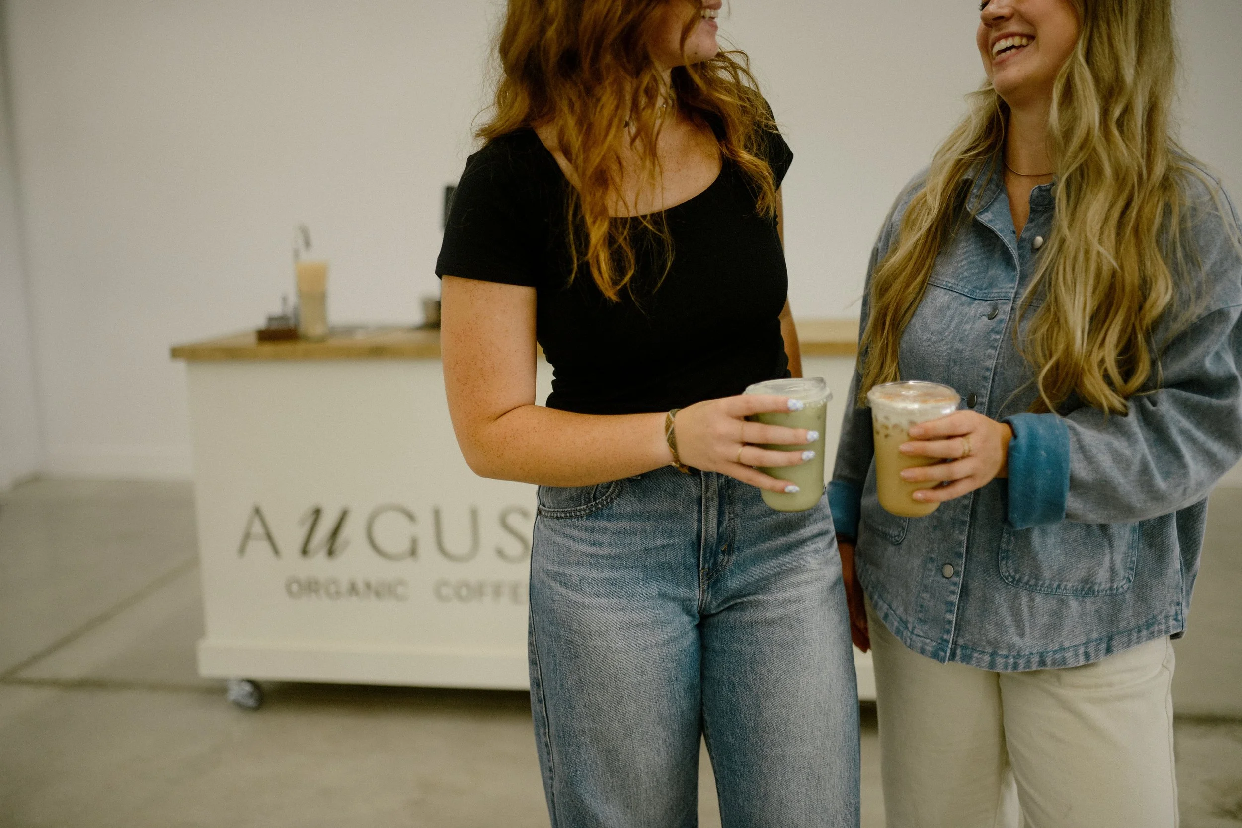 Two women smiling and holding drinks at a coffee shop has a sign that reads 'AUGUS'. They are wearing casual clothing, including denim and black tops.