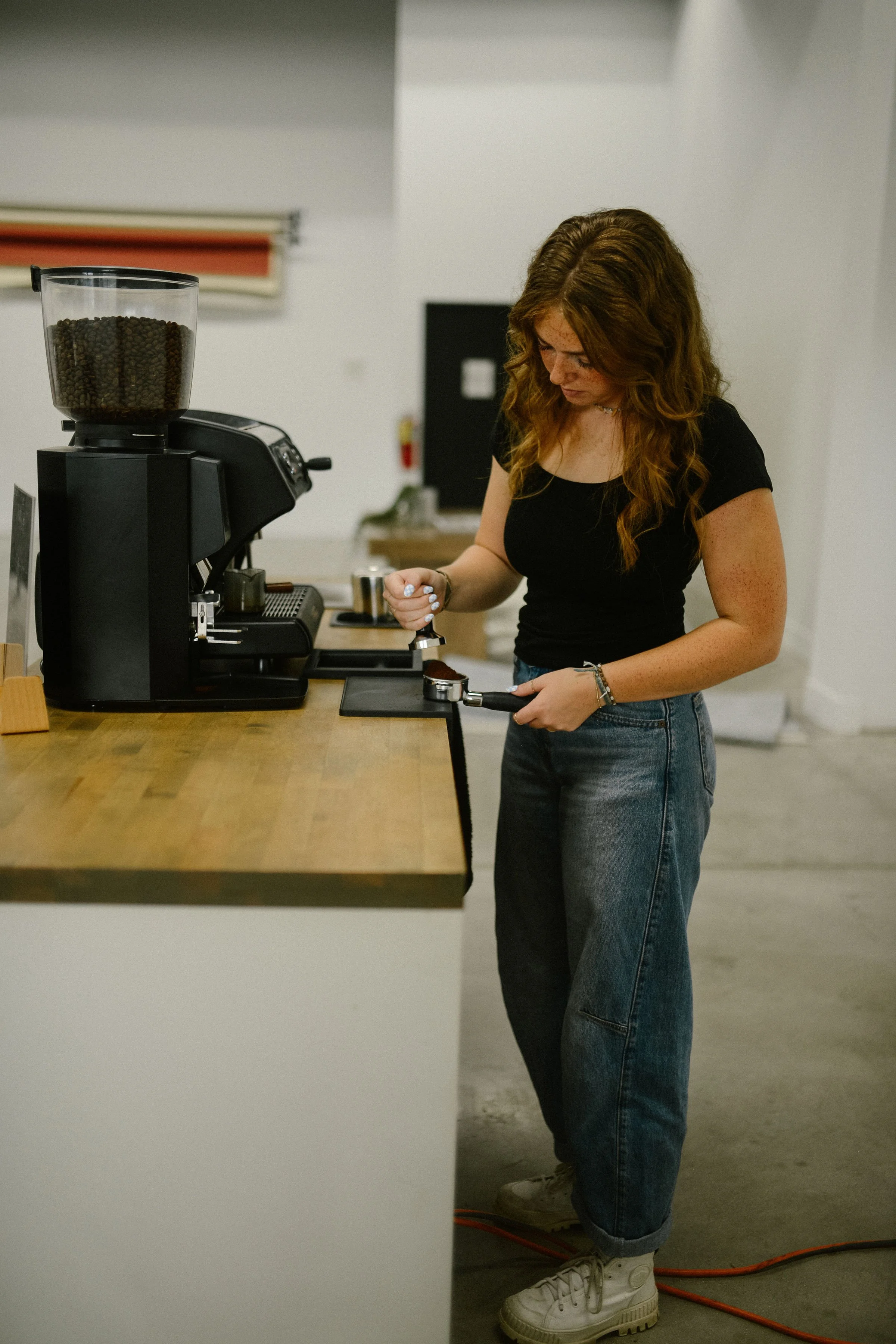A young woman with long, curly red hair wearing a black t-shirt, blue jeans, and white sneakers, standing in a room with white walls, using a portafilter to make espresso, with a coffee grinder filled with coffee beans on the countertop beside her.