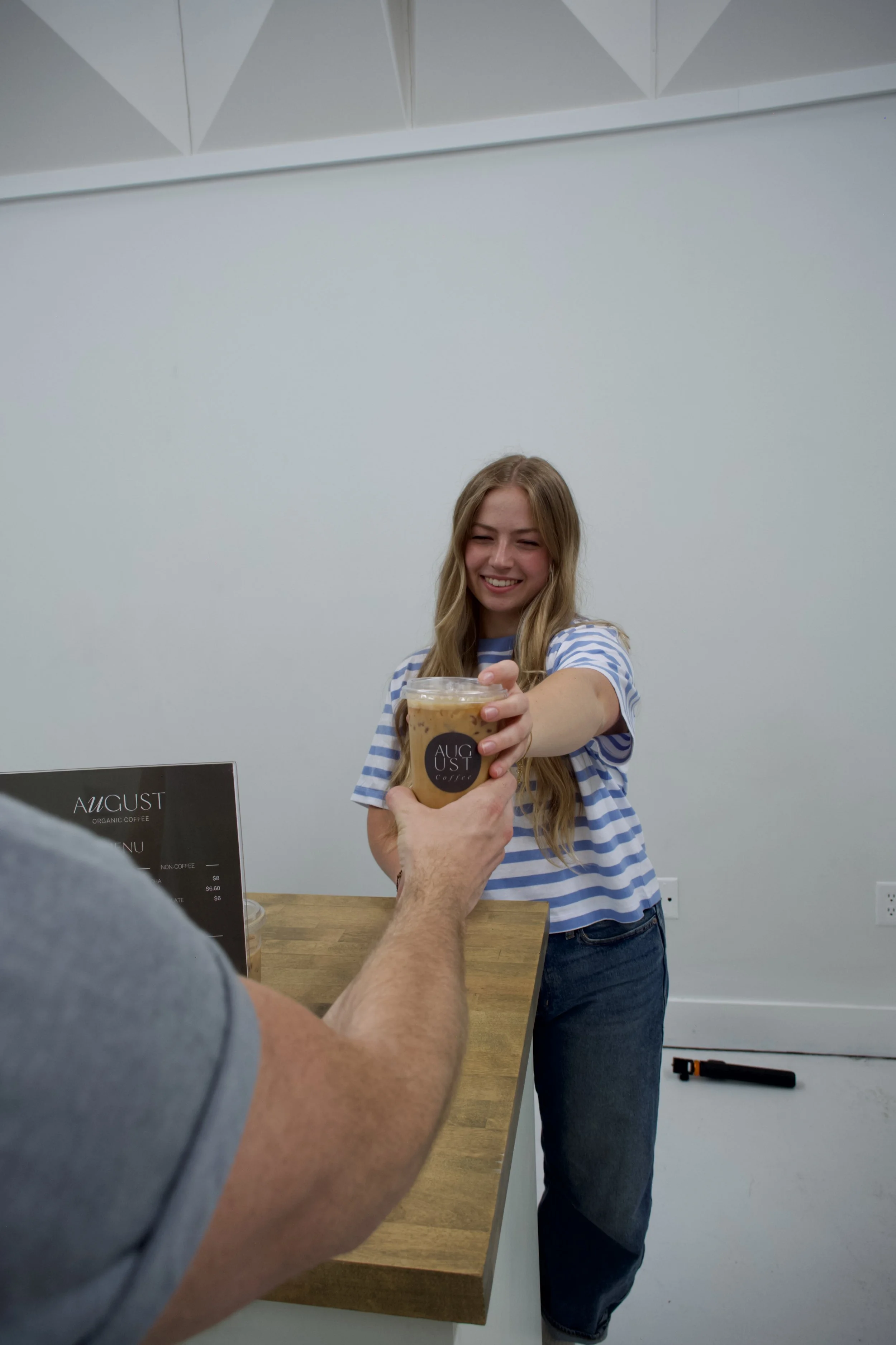 A woman with long blonde hair, wearing a striped shirt, smiling as she receives a coffee drink from someone at a counter. The person handing the coffee is partially visible, wearing a gray shirt. The background is a plain, light-colored wall with a menu board and some equipment on the wall.