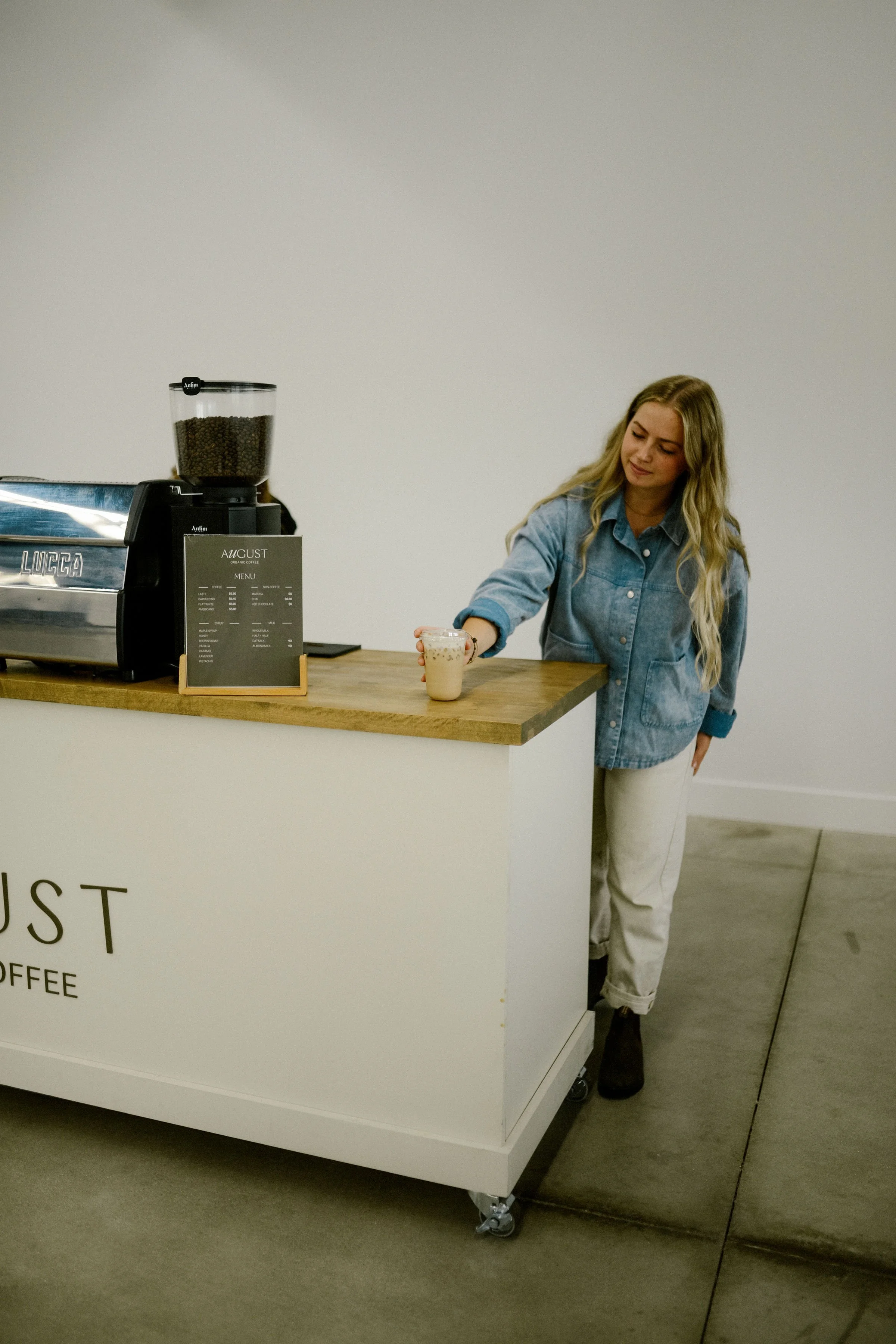 A woman with long blonde hair wearing a blue denim jacket and light-colored pants is standing at a coffee counter, handing a cup to a customer. The counter has a coffee machine, a grinder, and a menu sign. The background is a plain, white wall.
