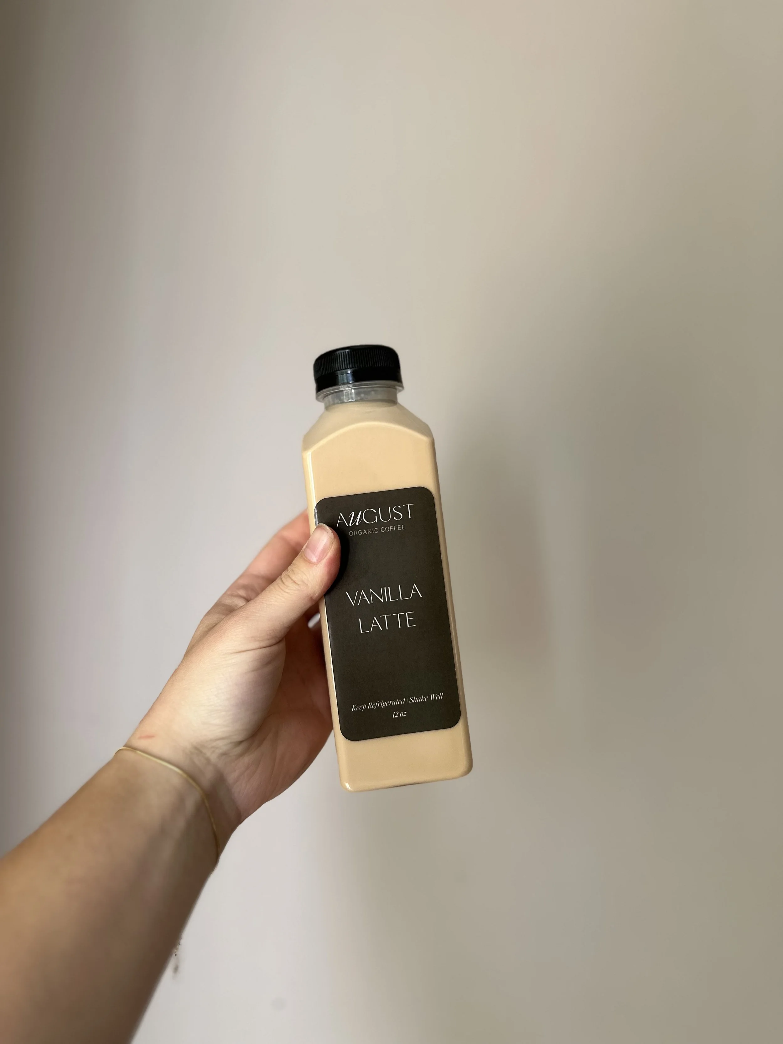 A hand holding a bottle of vanilla latte coffee labeled 'August Organic Coffee' against a plain wall background.