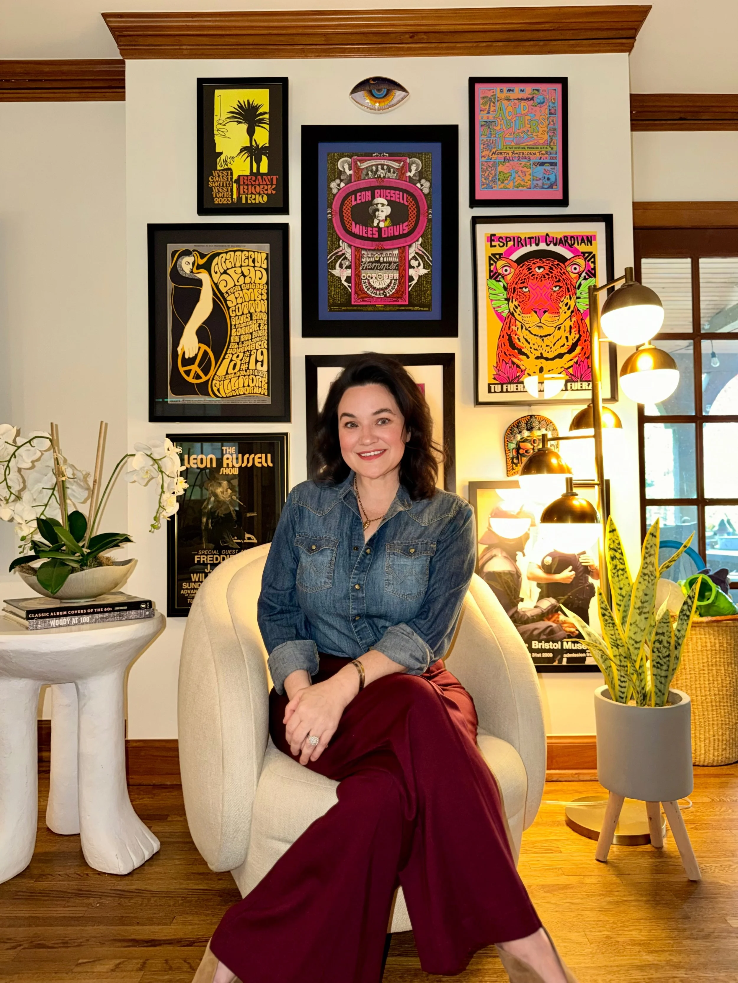 A woman with dark hair sitting in a cream-colored armchair in a room decorated with framed artwork on the wall, a potted plant, a white sculpture table with books, and a standing lamp with gold-colored bulbs. She is wearing a denim jacket and maroon pants and smiling at the camera.