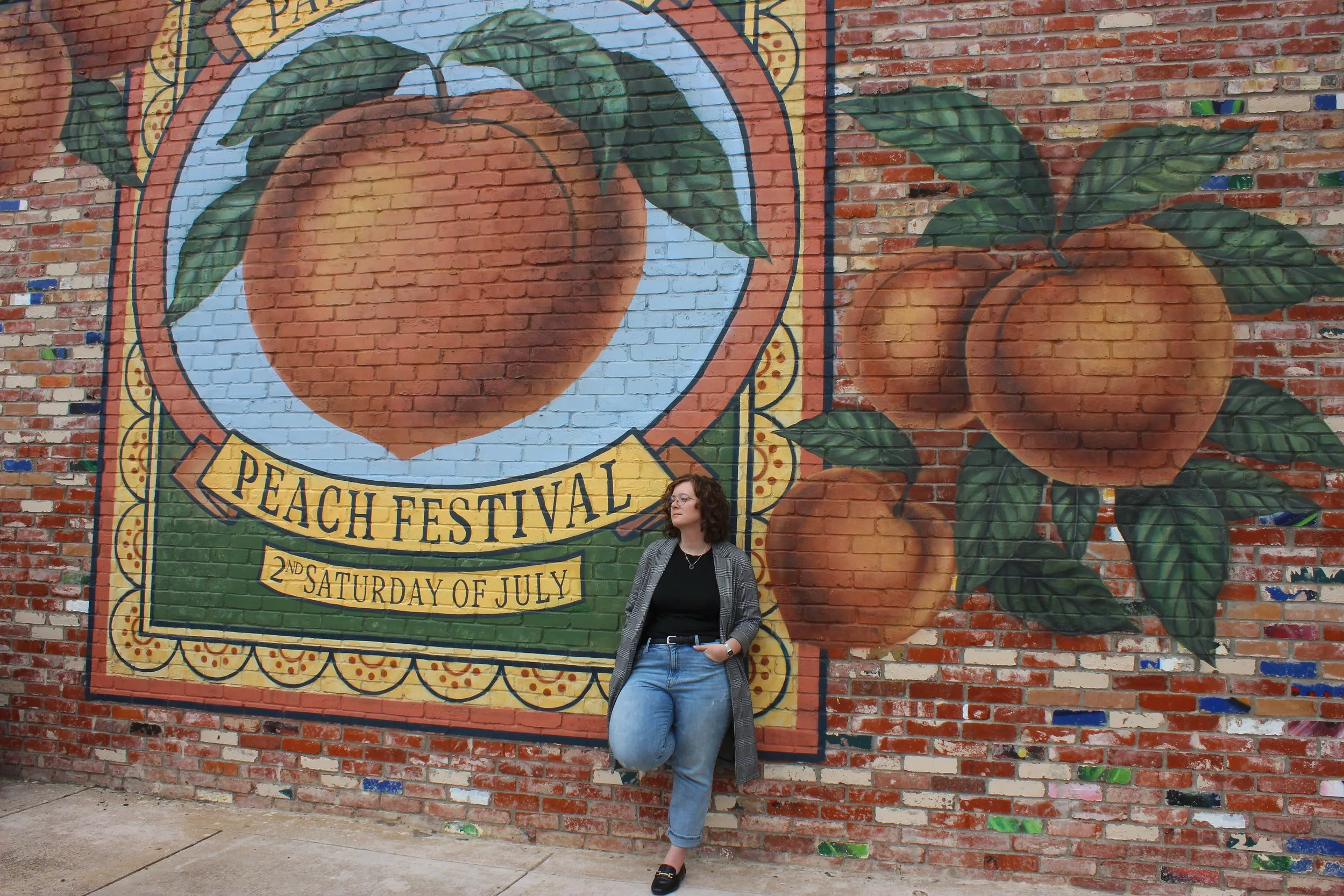 Kalli Stout standing beside a brick mural celebrating the Weatherford Peach Festival.