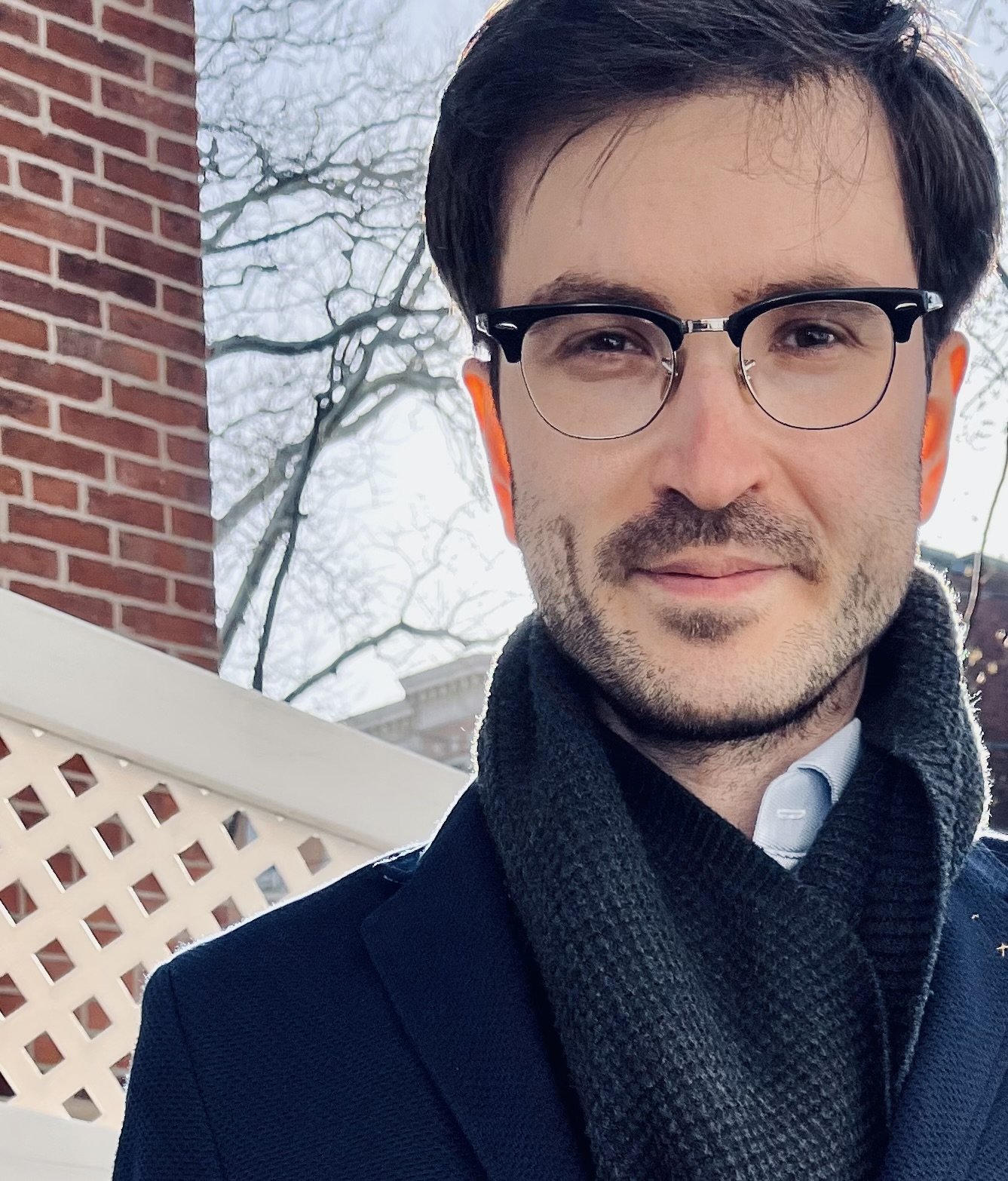 Tomas Baka, physician-scientist; man with dark hair, glasses, and a light beard, wearing a dark coat and a scarf, standing outdoors in front of a white lattice fence, trees, and a red brick building.