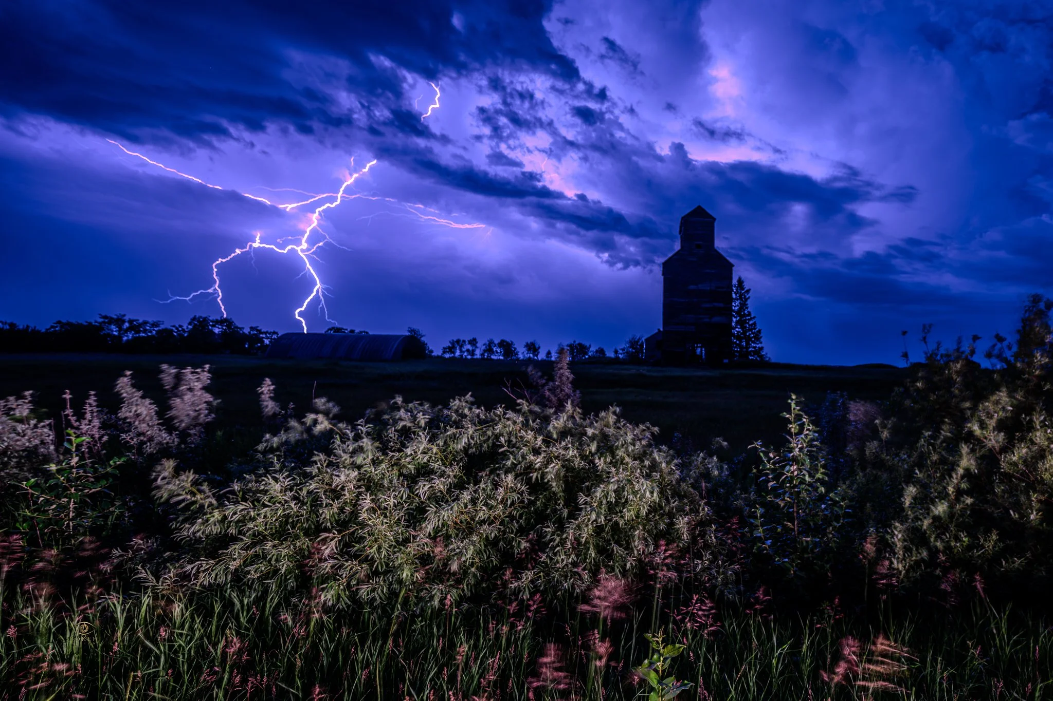 Nighttime scene with dark sky filled with purple and blue clouds, visible lightning strikes, a silhouette of a building with a tower, farm structures, and lavender flowers in the foreground.
