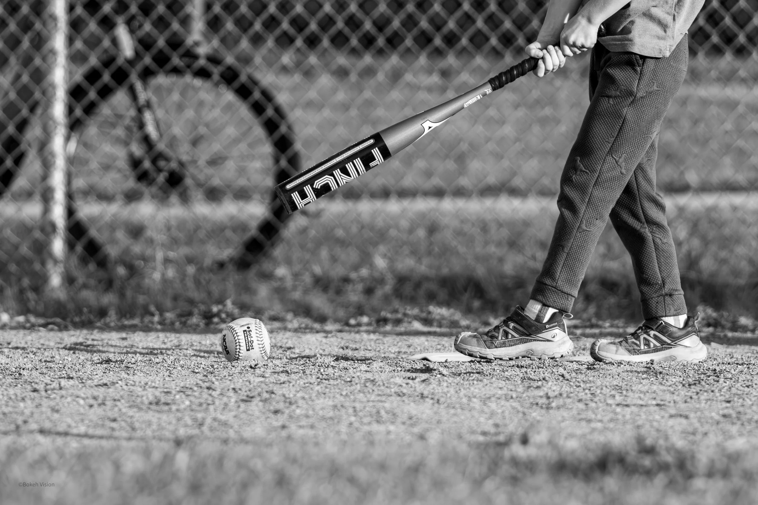 Child in casual sportswear prepares to hit a baseball with a bat on a baseball field, with a baseball on the ground nearby and a chain-link fence in the background.