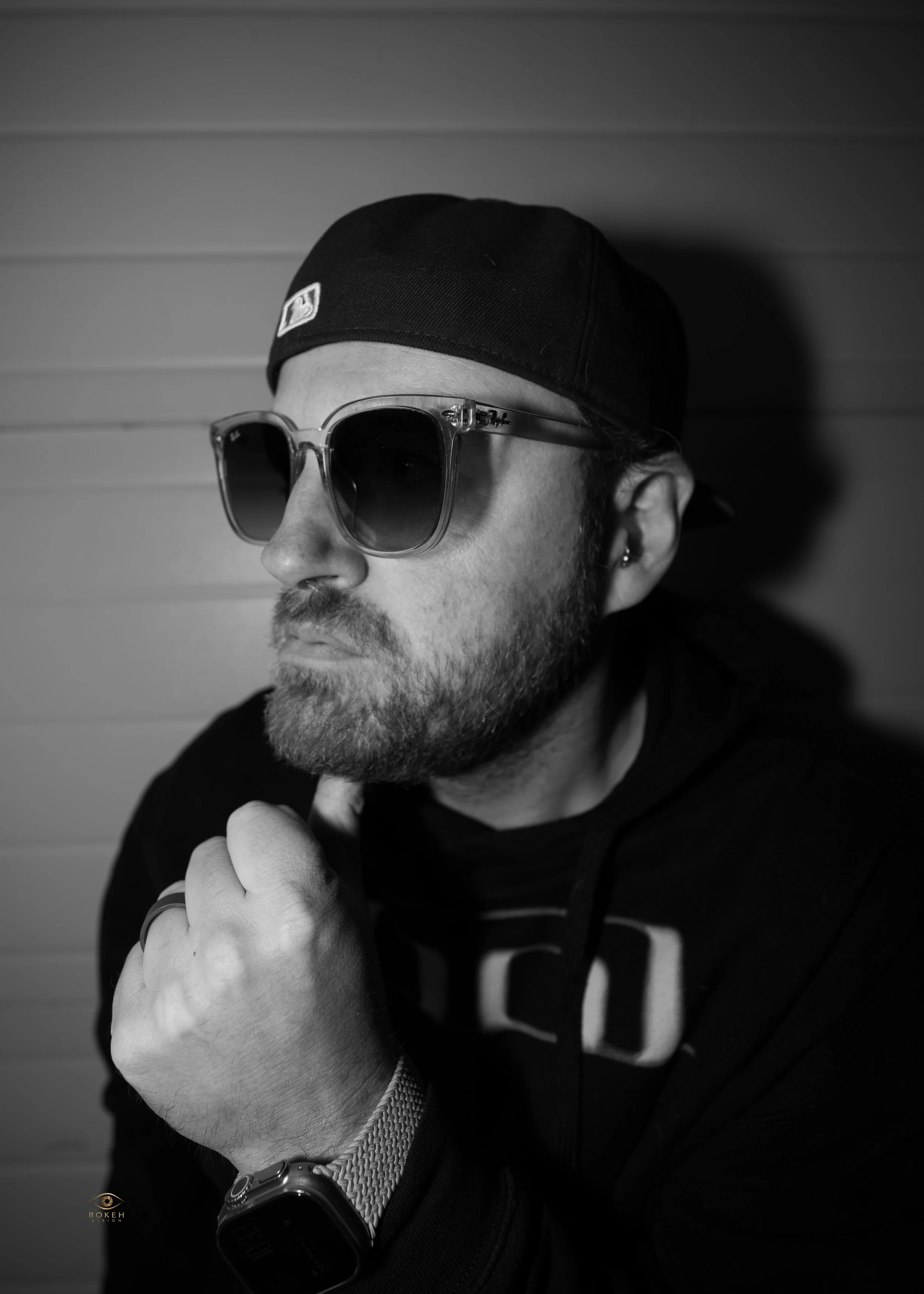 Black-and-white photo of a man wearing sunglasses, a black cap, a hoodie, and a watch, with a background of horizontal wood paneling.