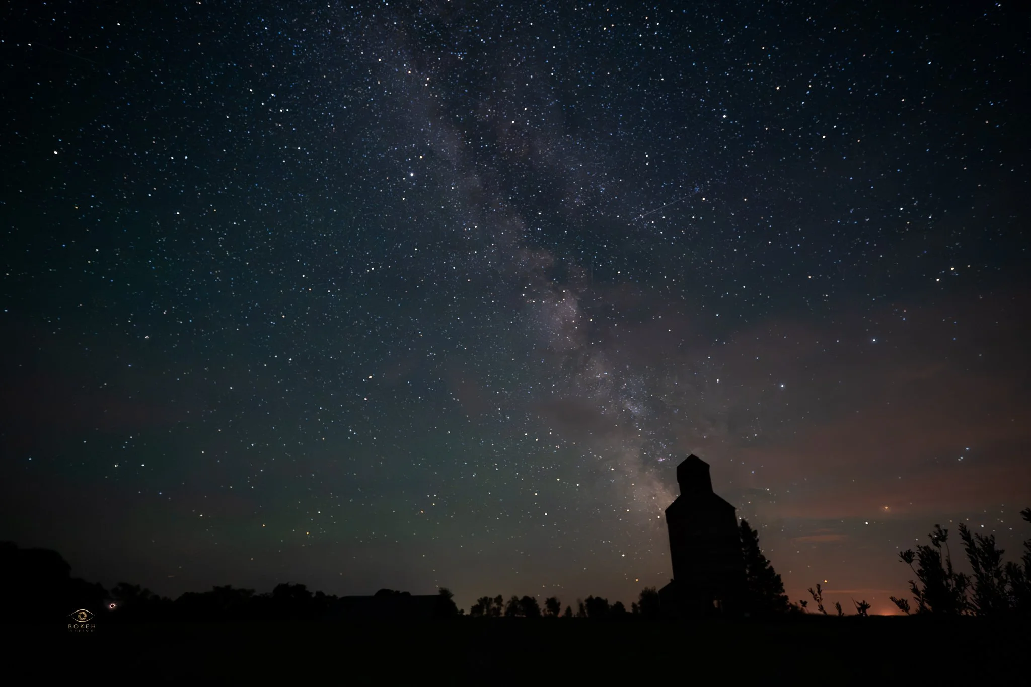 Night sky filled with stars and the Milky Way galaxy, with silhouettes of trees and a small building in the foreground.