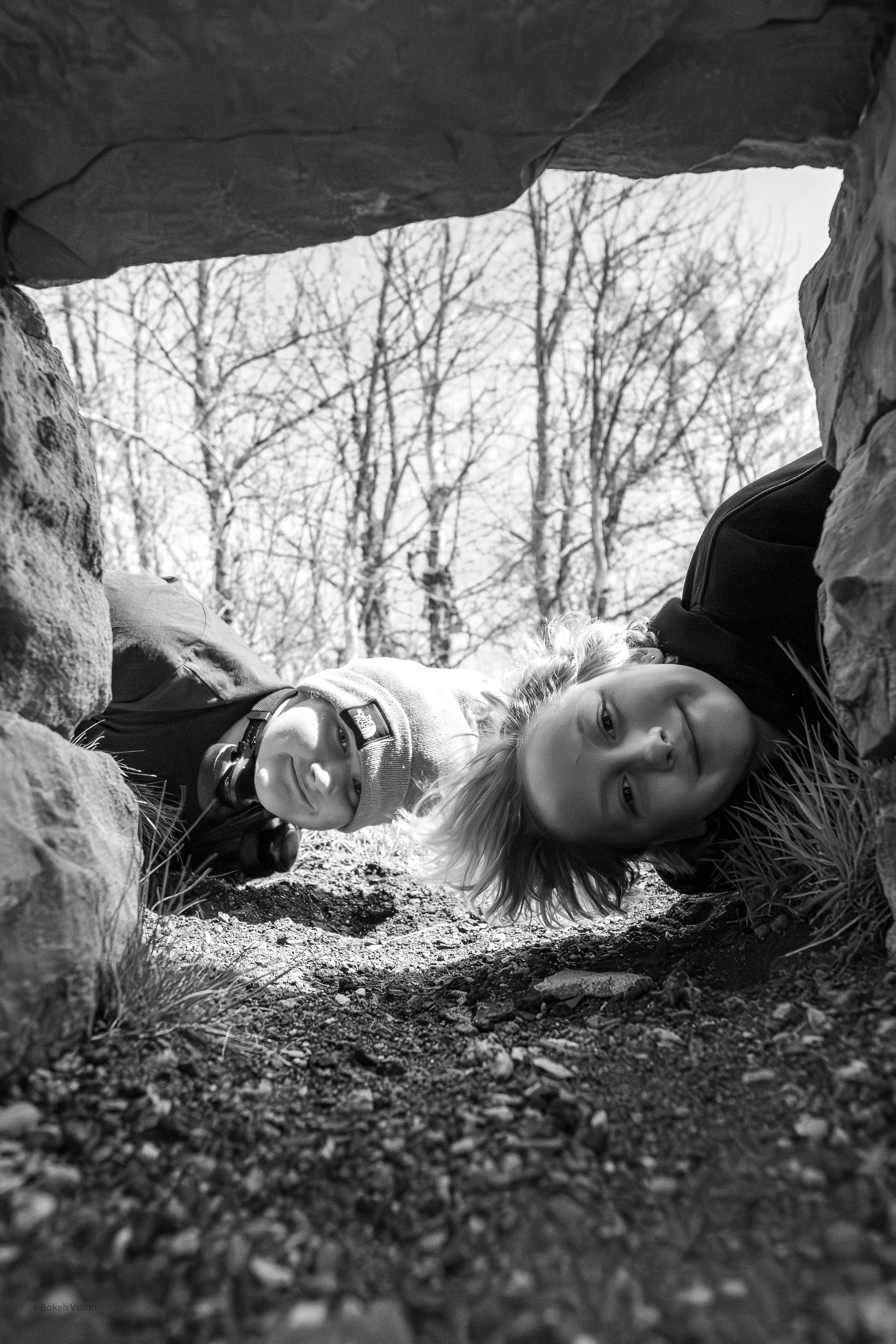Two children lying on the ground under a stone structure, looking at the camera; one wears glasses, and both have light-colored hair; trees with leafless branches are visible in the background.