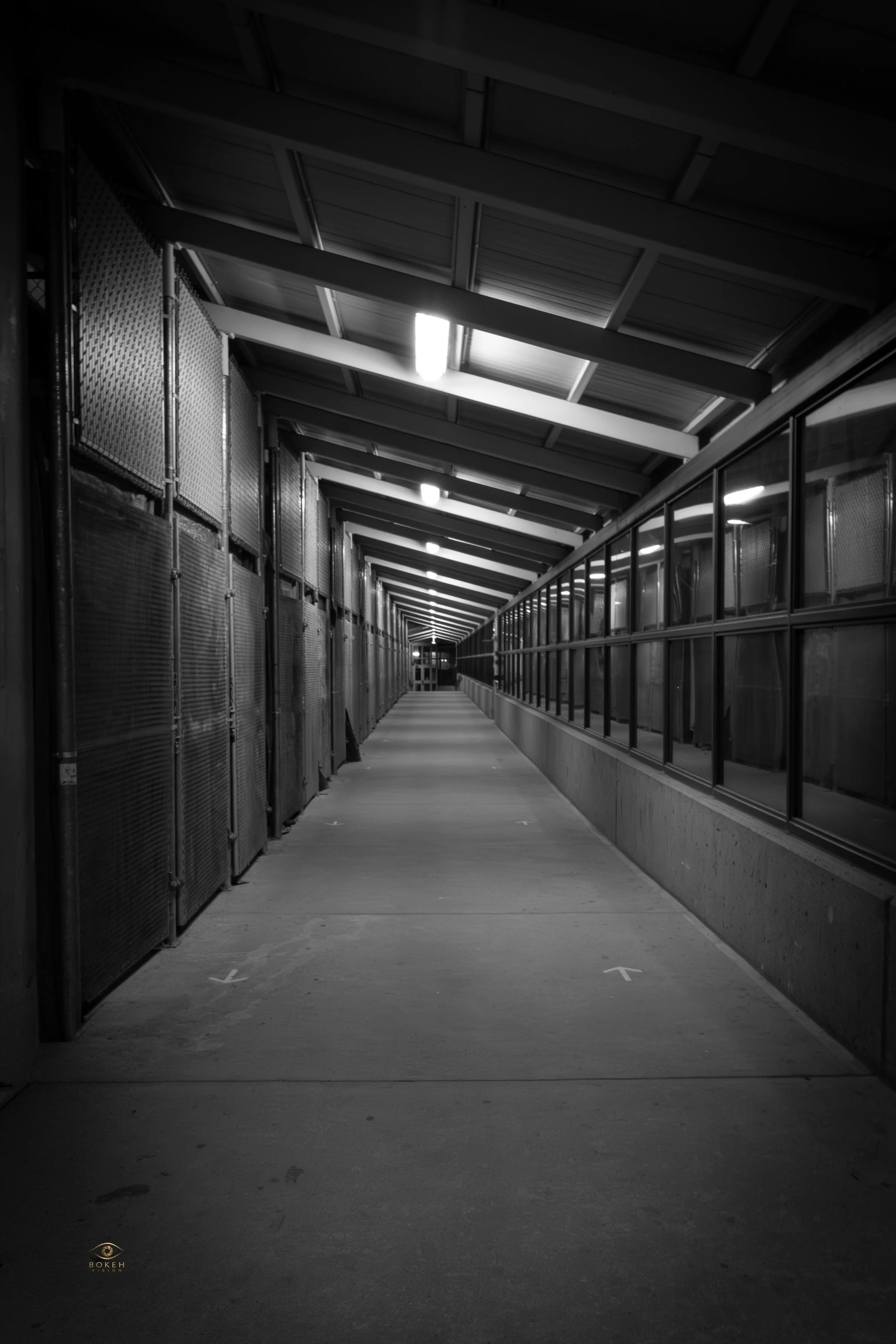 A long corridor with metal fences on the left and glass panels on the right, illuminated by bright overhead lighting, with a concrete floor and a ceiling with structural beams.